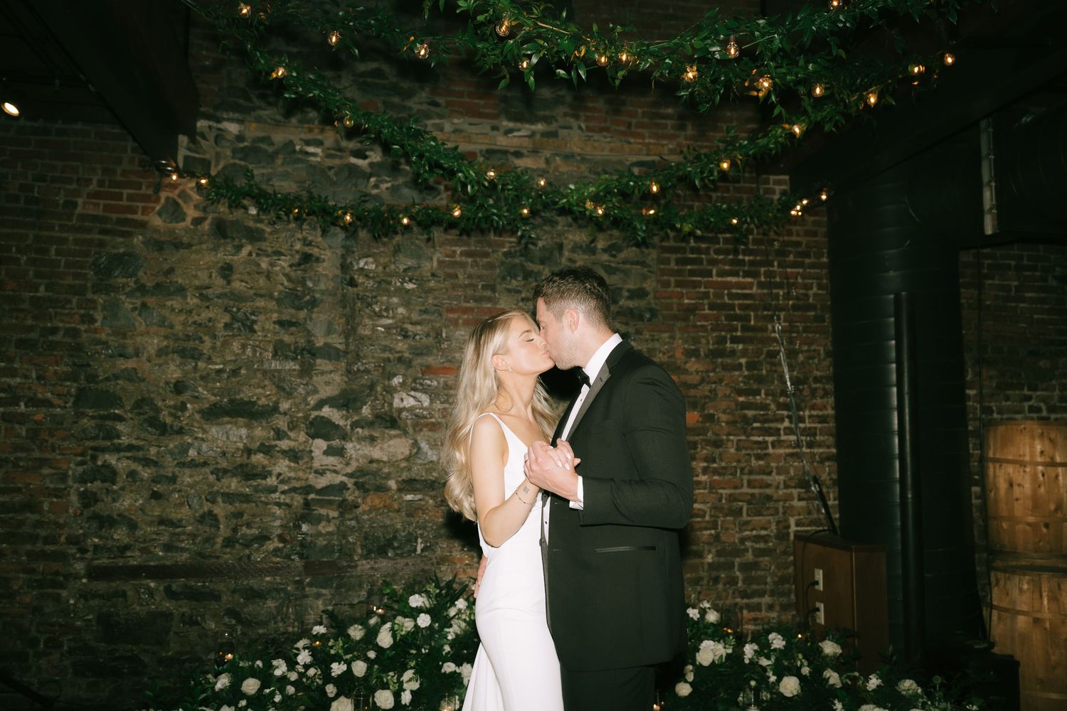 A romantic wedding setting with string lights and exposed brick walls decorated with greenery.