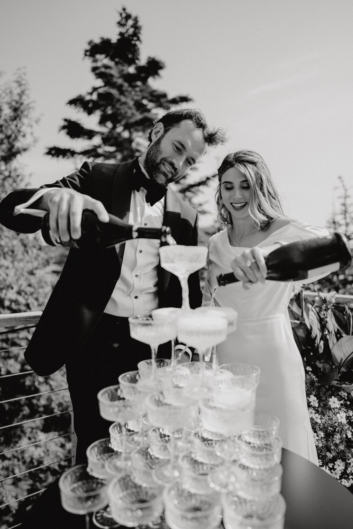 Guests pour champagne into a tower of elegant crystal glasses at an outdoor reception.