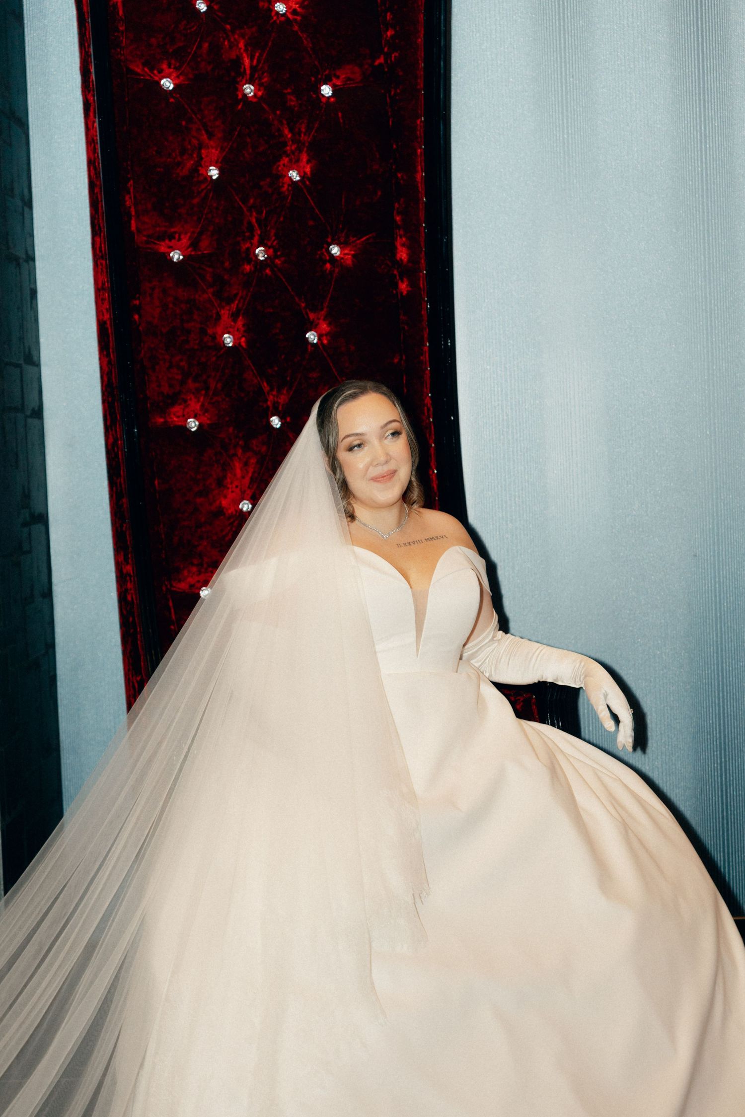 A bride wearing a voluminous white veil posing against a red sparkly backdrop.