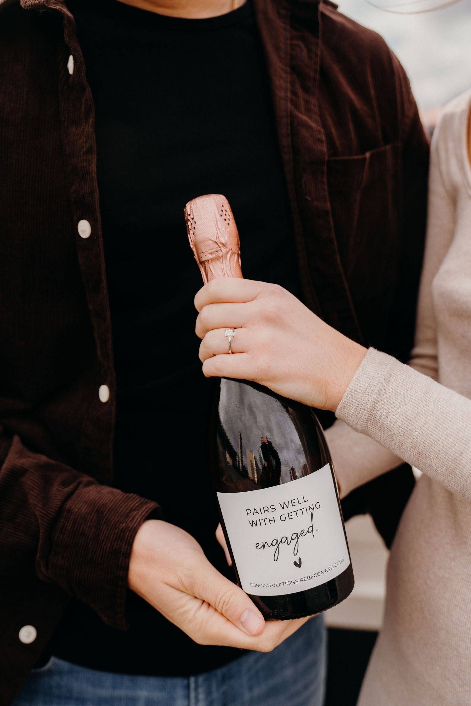 Two people holding a bottle of champagne labeled 'engaged' during a rooftop celebration.