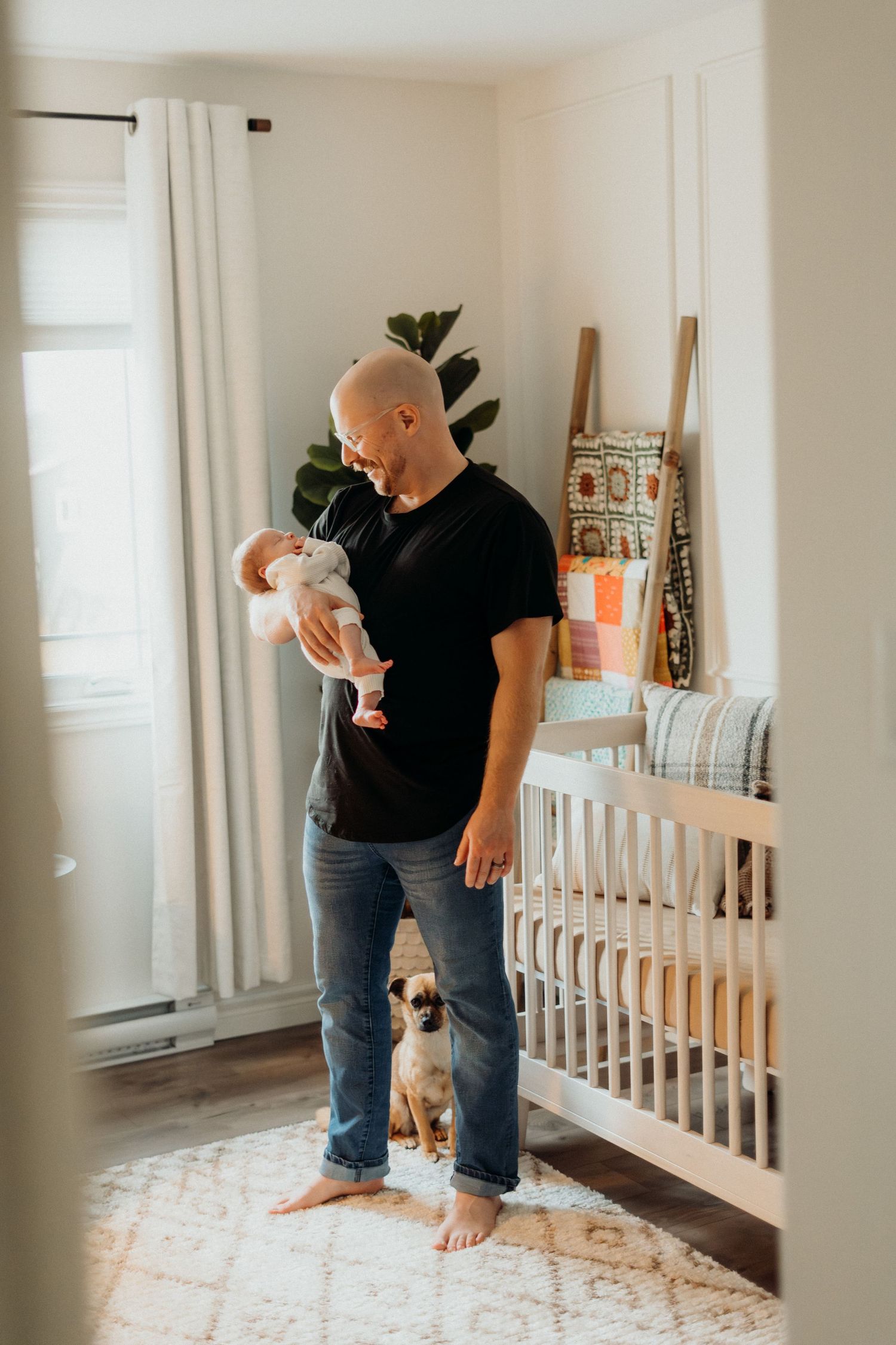 Caregiver in black shirt interacting with baby near white crib and windows.