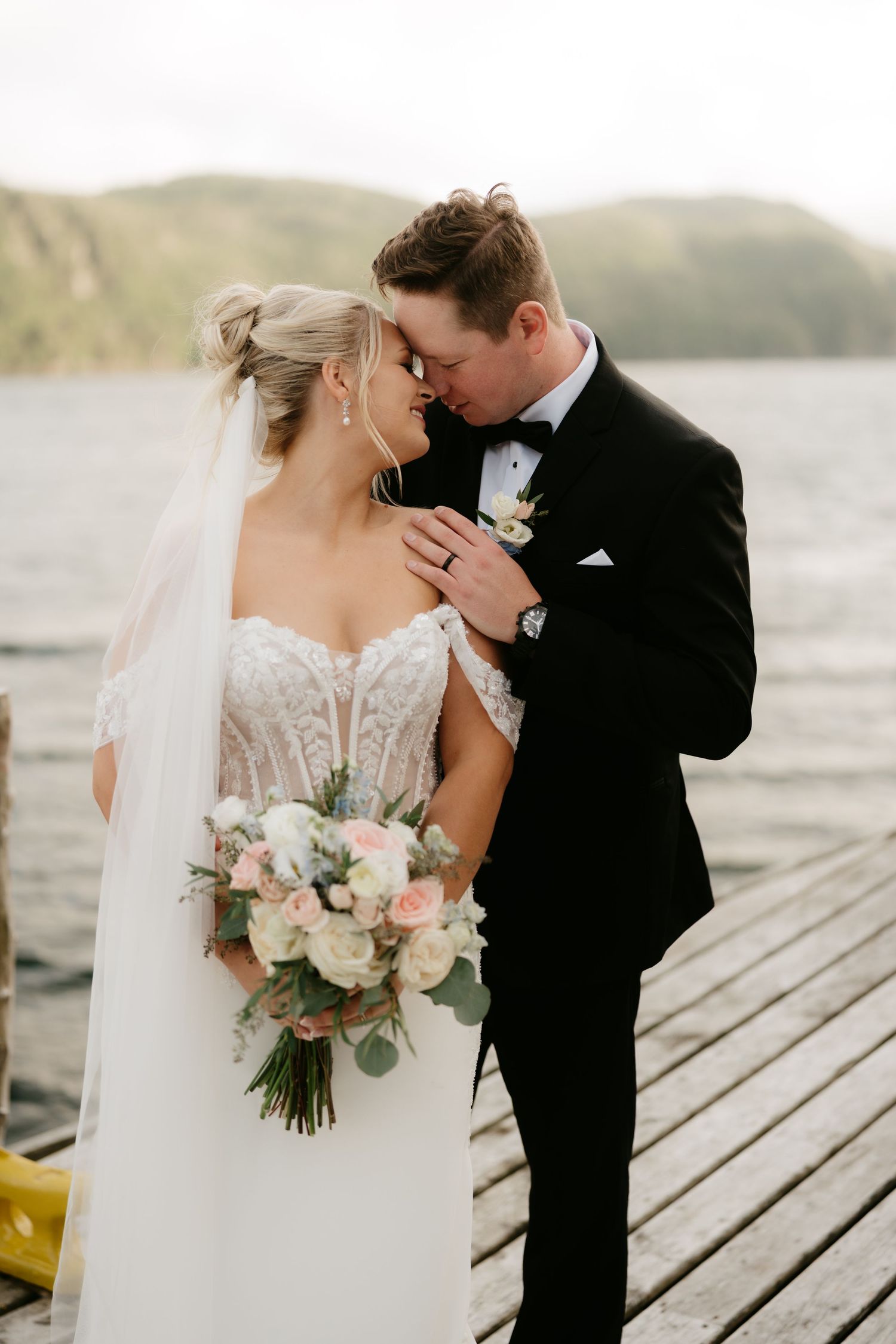 Bride and groom share a romantic moment on a wooden dock, with mountains and water in the background.