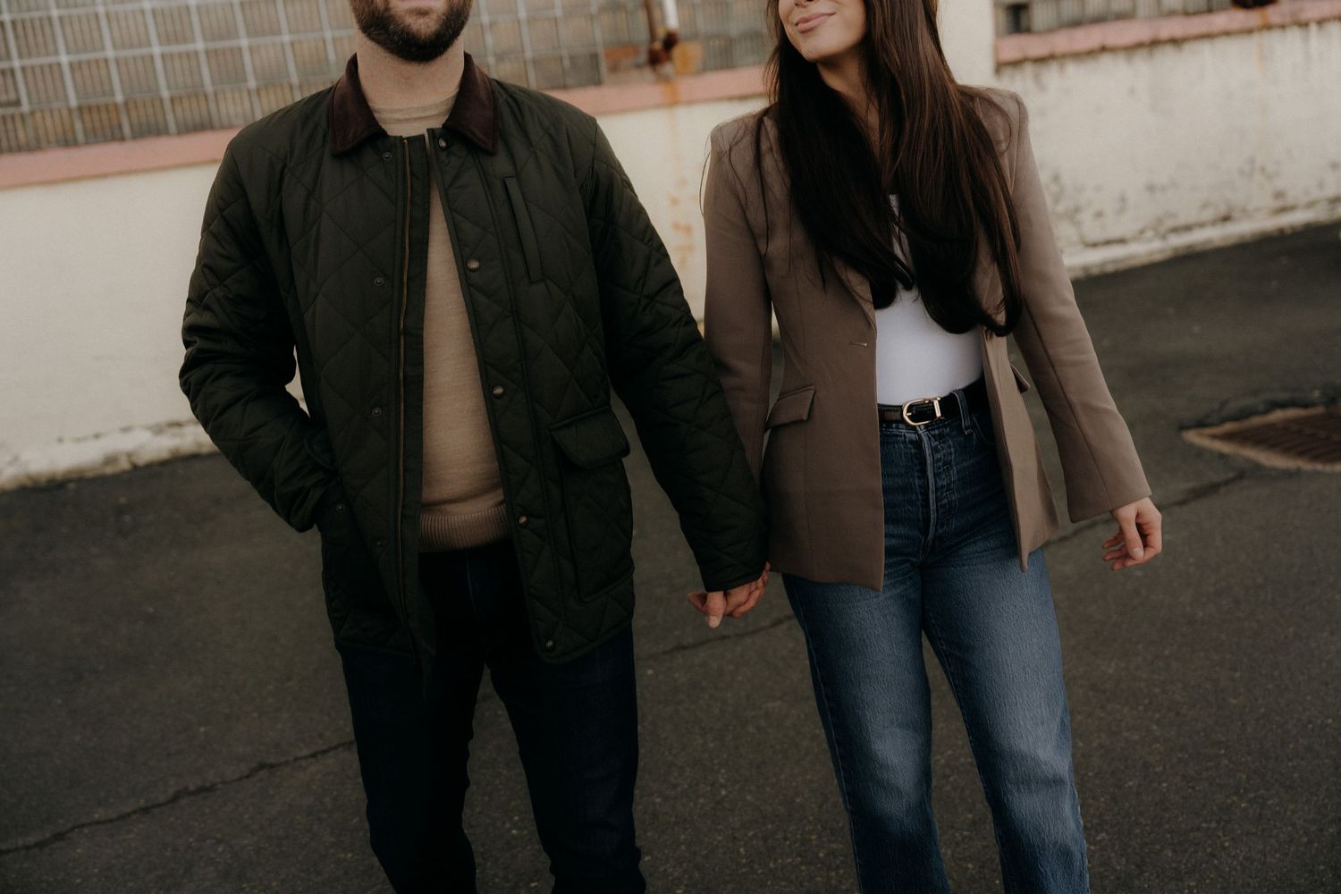 Couple holding hands outdoors, man in dark jacket and woman in brown blazer with jeans, urban setting.