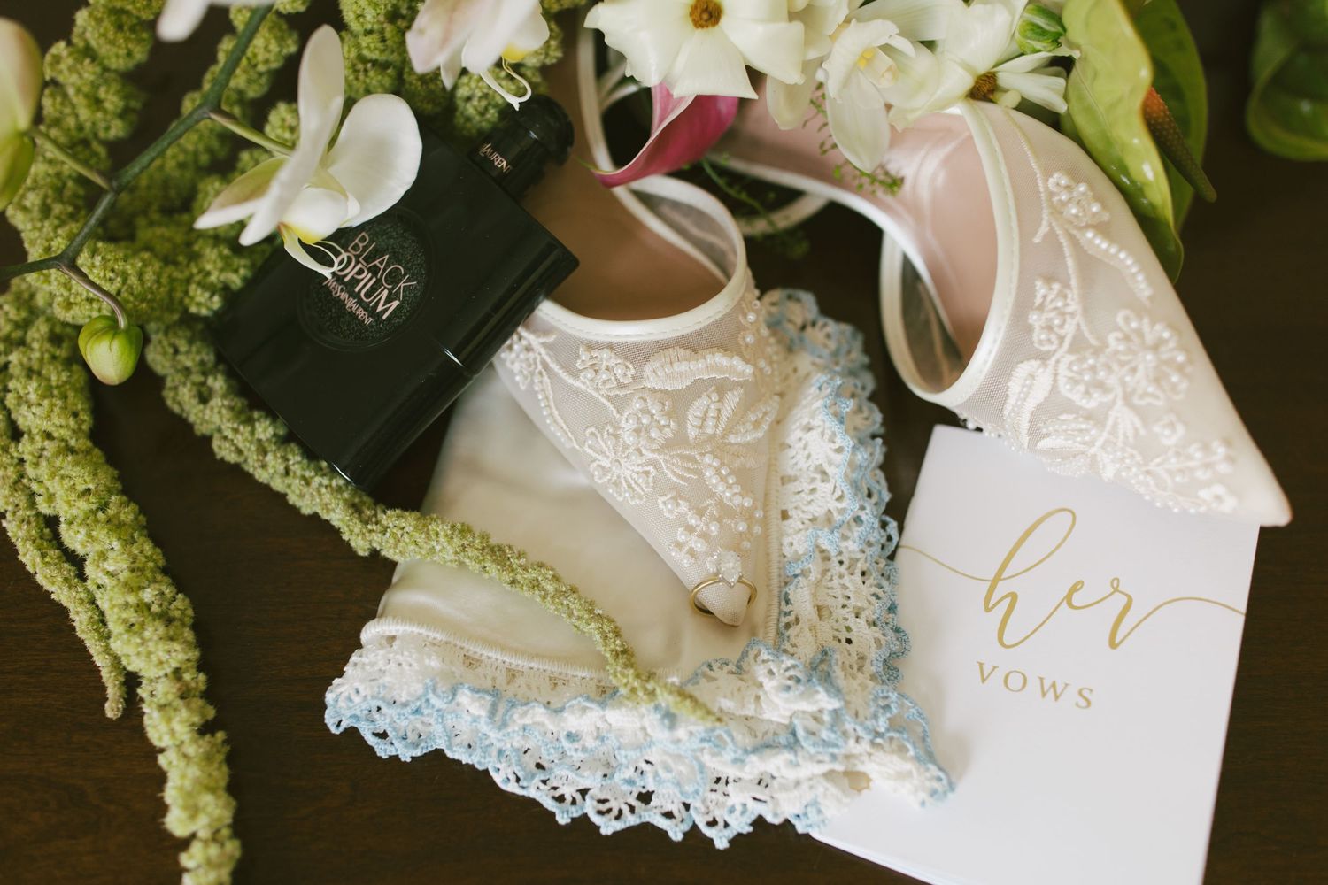 Bridal flat shoes with lace detail beside her vows card, bouquet, and blue garter on wooden surface.