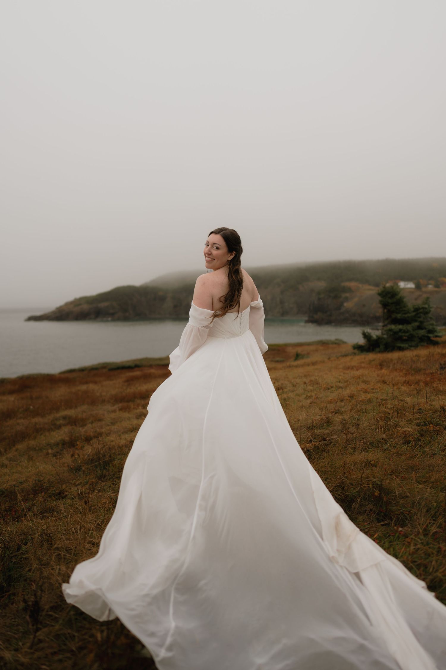 A flowing white wedding dress billows dramatically against a misty coastal landscape on a moody afternoon.