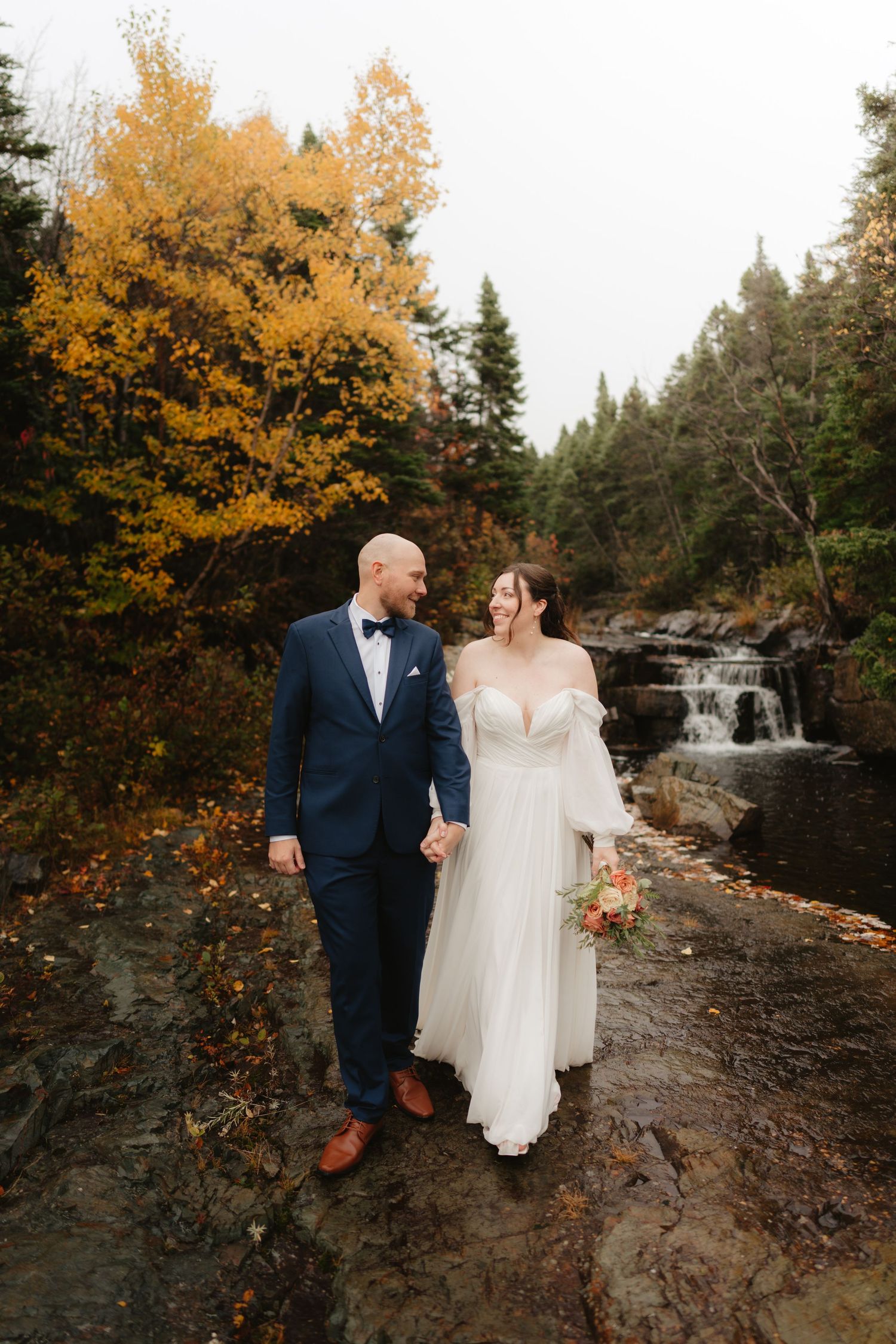 A couple in wedding attire stands on rocky terrain near a waterfall surrounded by autumn foliage in a forest setting.