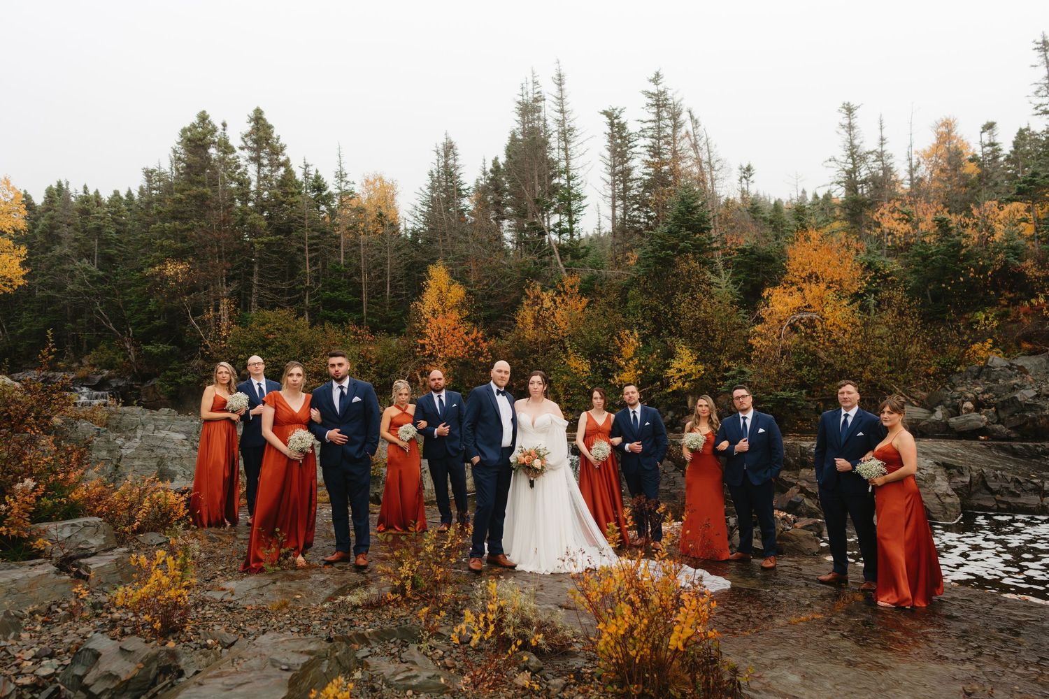 Wedding party stands together on rocky terrain surrounded by autumn trees and golden foliage at sunset.