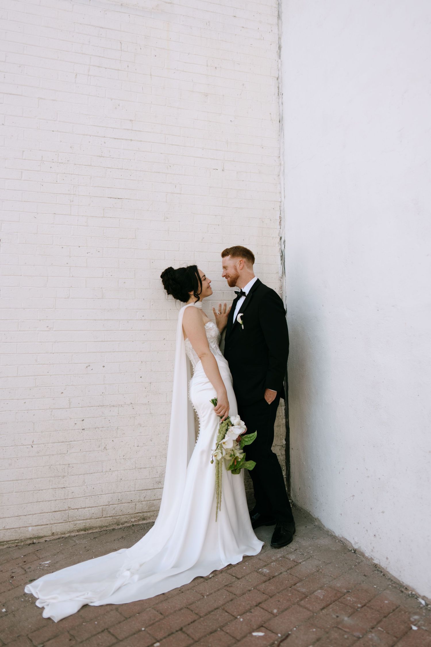 Bride in white gown with cape and groom in black tux share a romantic moment against a white brick wall.