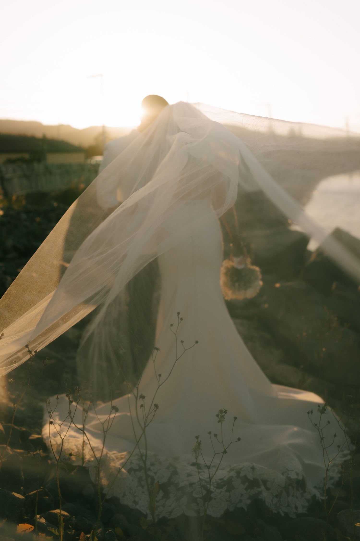 A flowing white wedding veil catches the golden sunlight against a dreamy outdoor landscape at sunset.