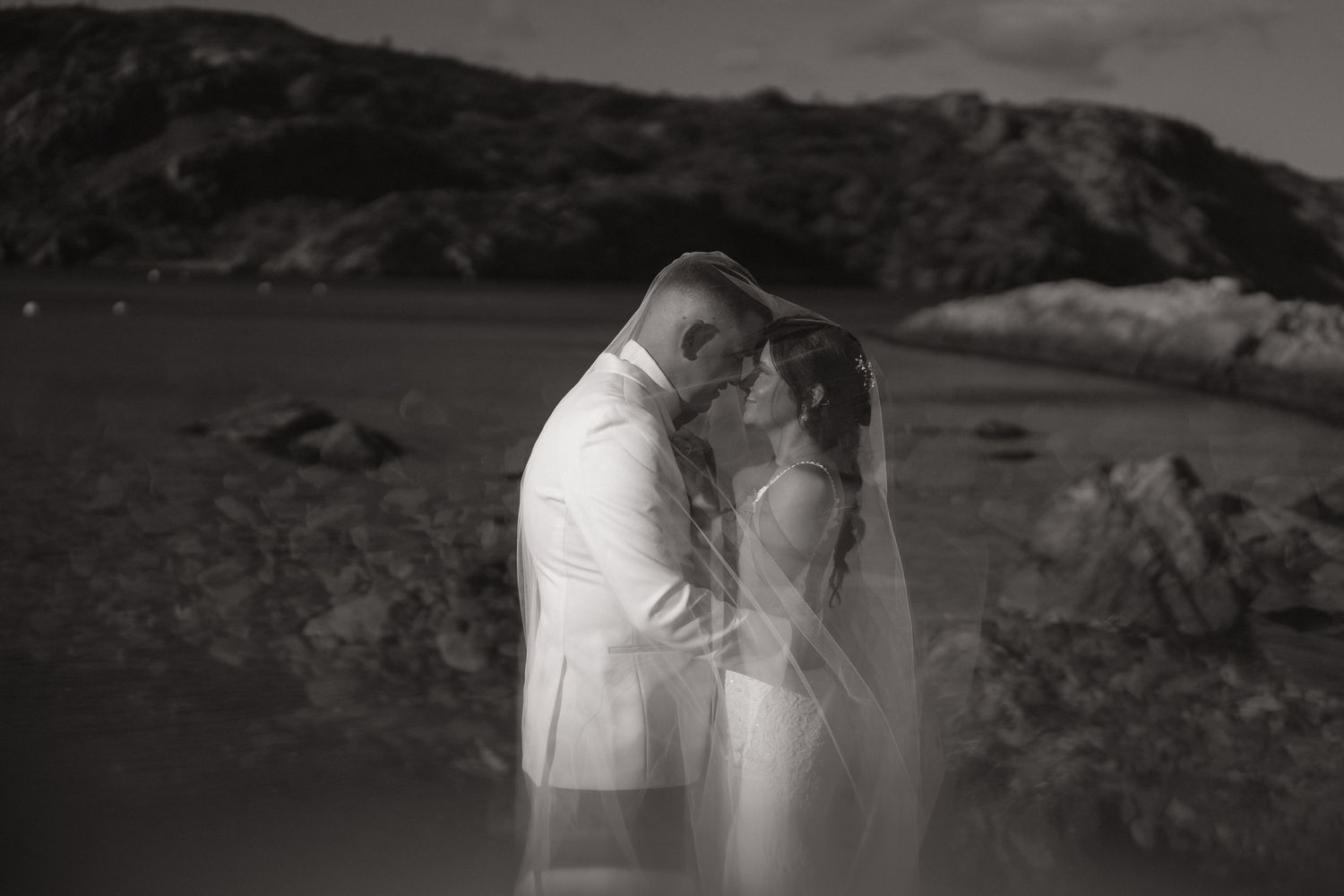 A romantic black and white photograph of a couple embracing on a rocky beach with mountains in the background.