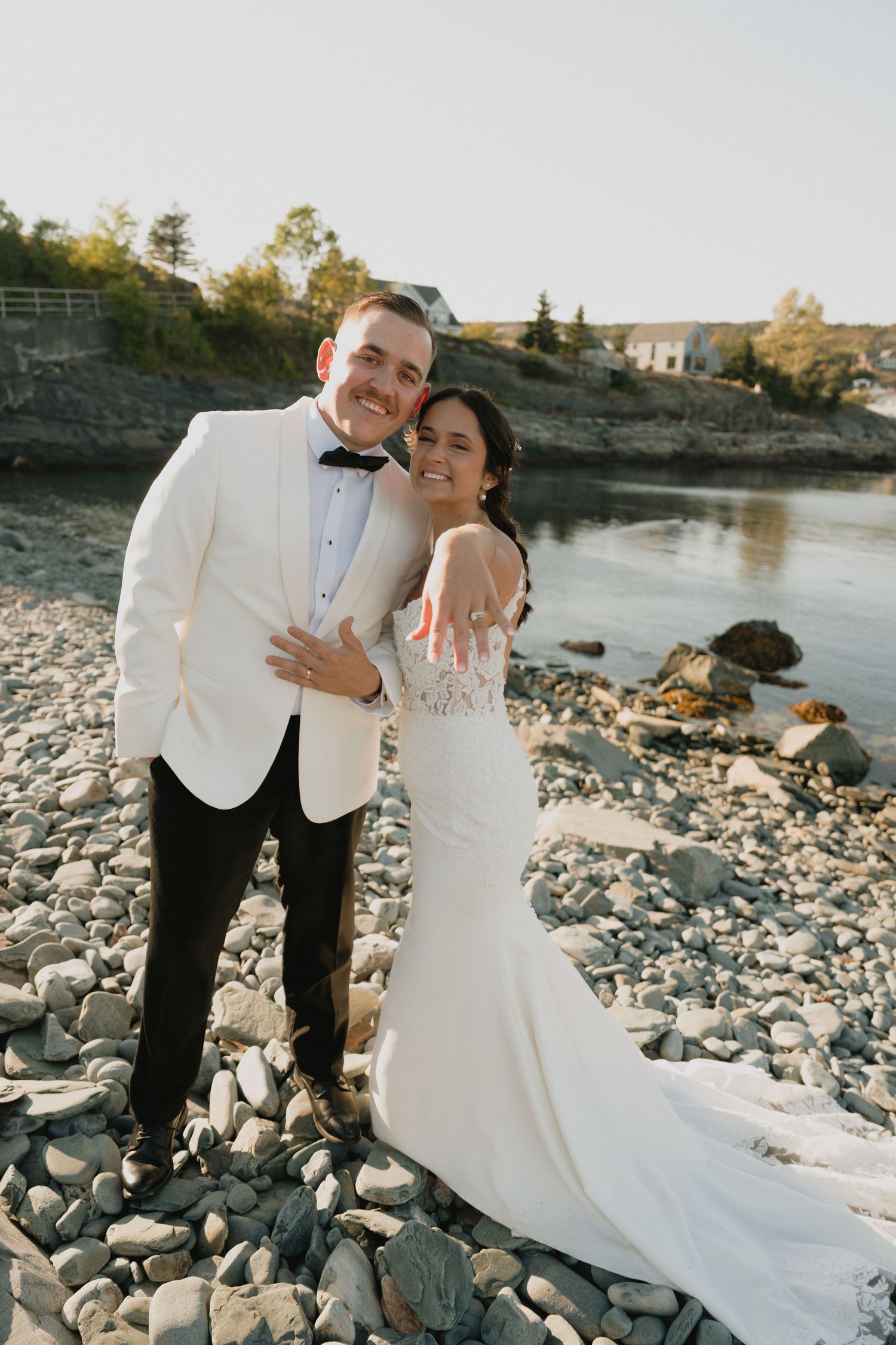 Romantic wedding couple embraces on rocky shoreline at sunset with water and houses in background.
