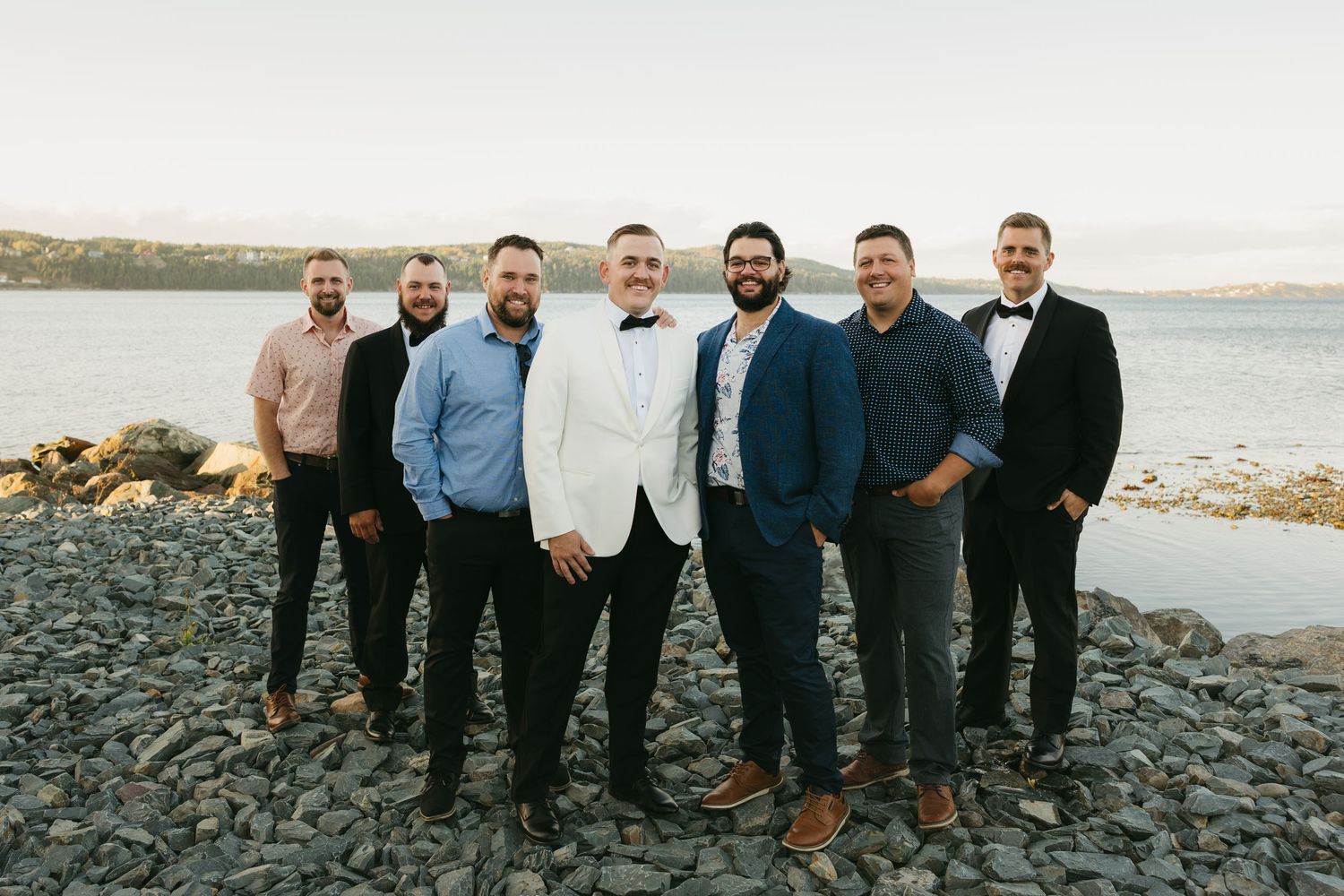 A group in formal attire stands together on a rocky beach shoreline during an outdoor wedding celebration.