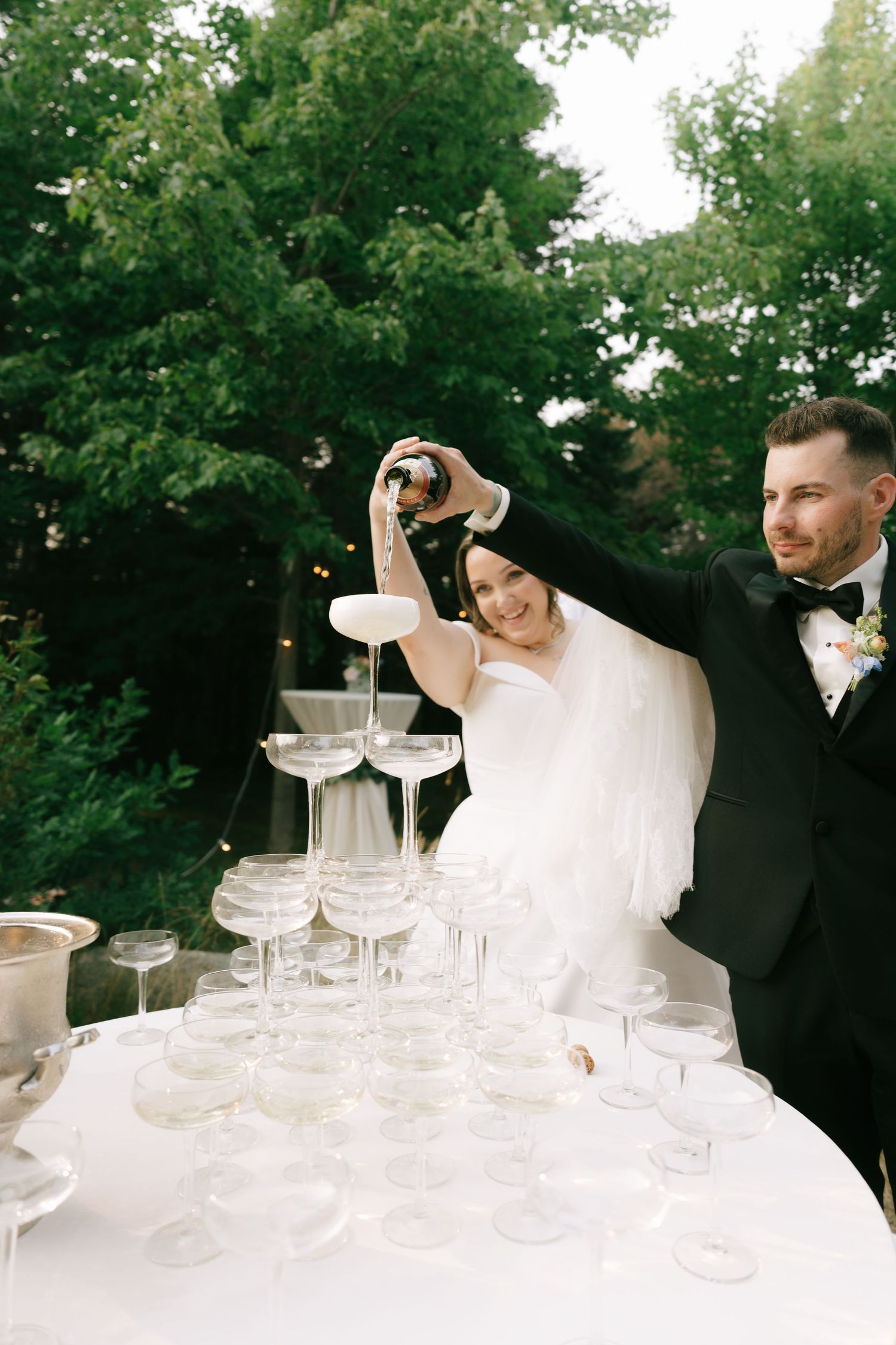 Champagne is being poured into an elegant tower of stacked crystal glasses at an outdoor wedding celebration.