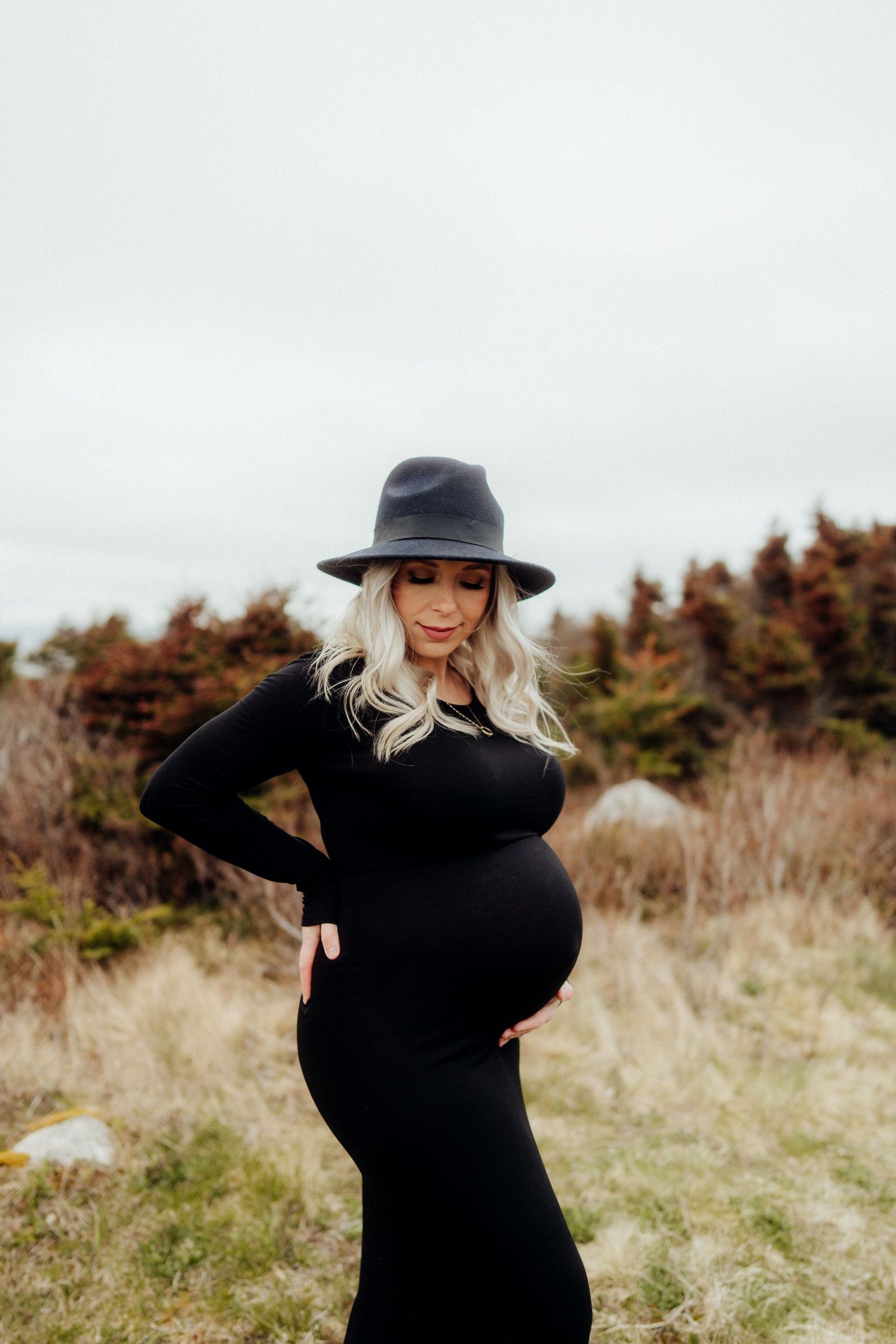 A person in black poses against an autumn landscape wearing a stylish hat.