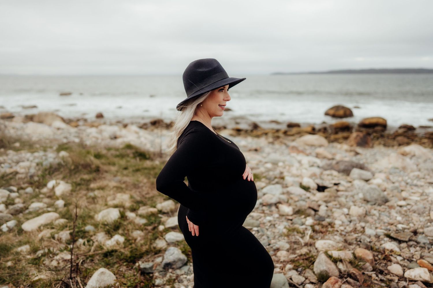 Silhouette of pregnant woman in hat against rocky coastal background.
