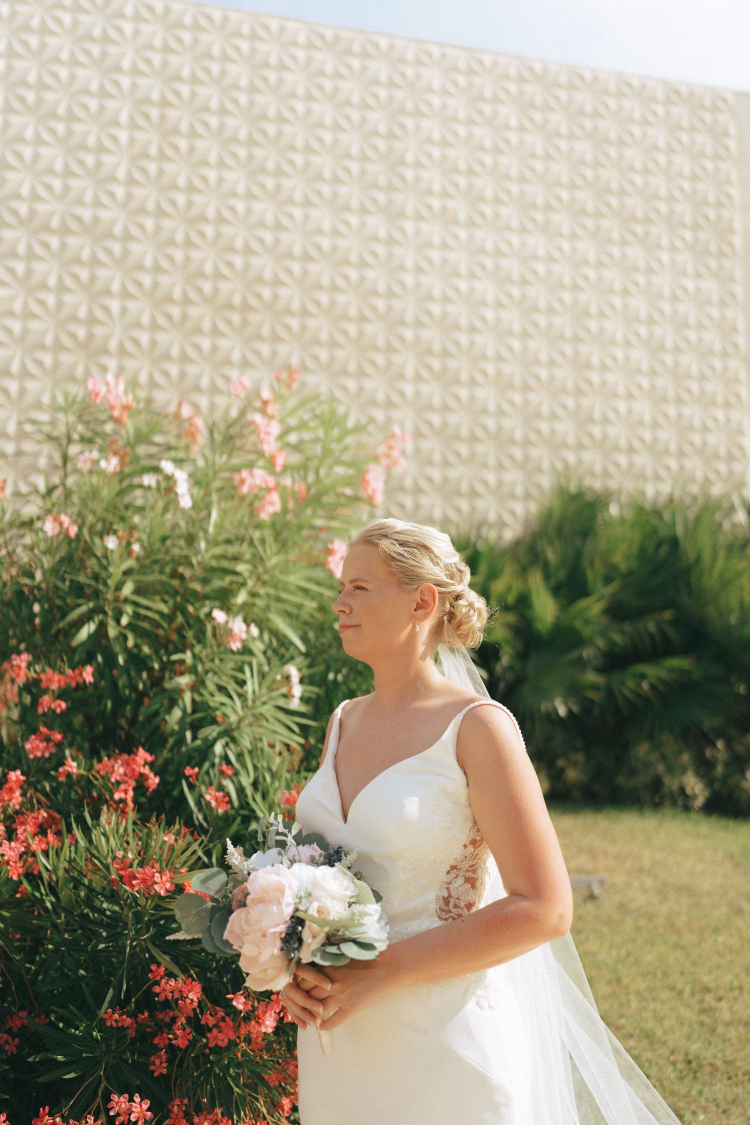 Bride in a white wedding dress holding a bouquet against a wall with coral flowers.