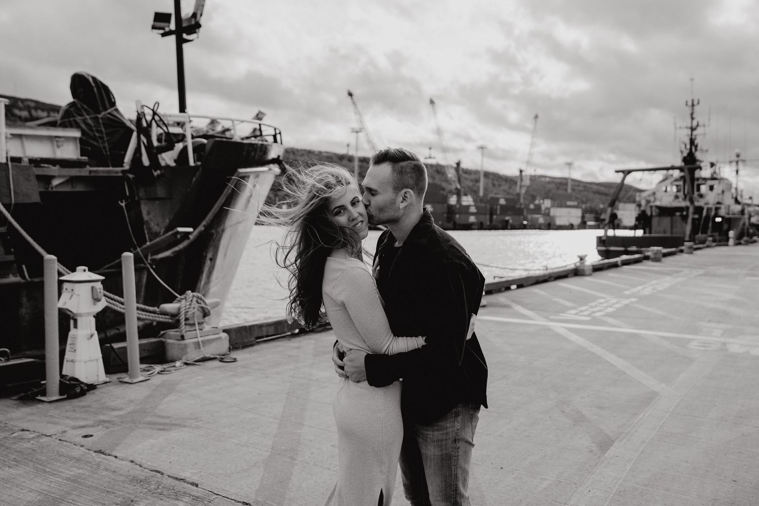 A romantic scene unfolds on a harbor dock with industrial ships and cloudy weather.