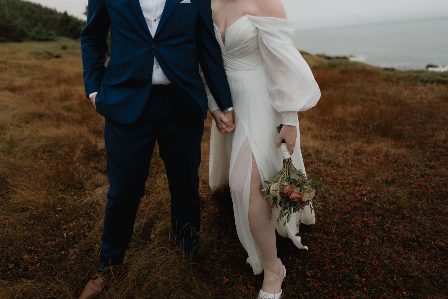 A couple in wedding attire walks together along a scenic coastal cliff during sunset.