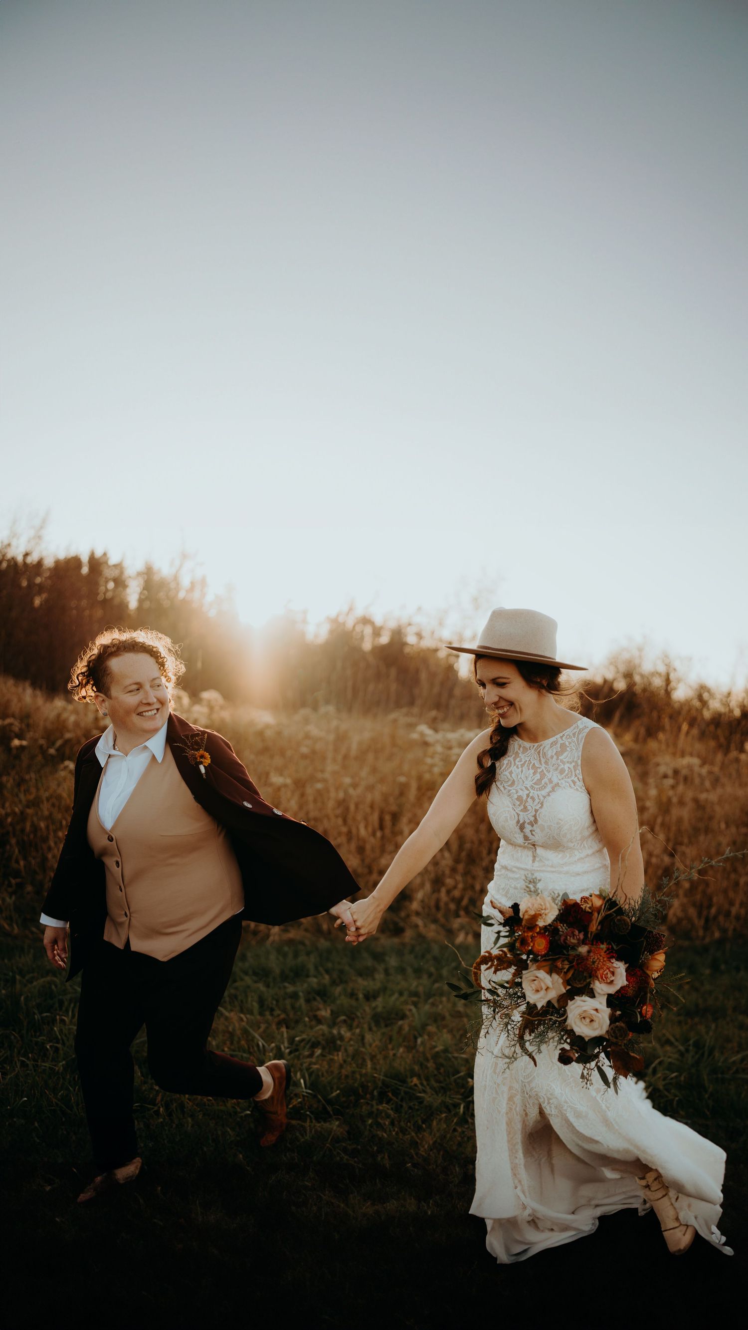 A couple in wedding attire holds hands and dances in a golden sunlit field at sunset during their outdoor celebration.