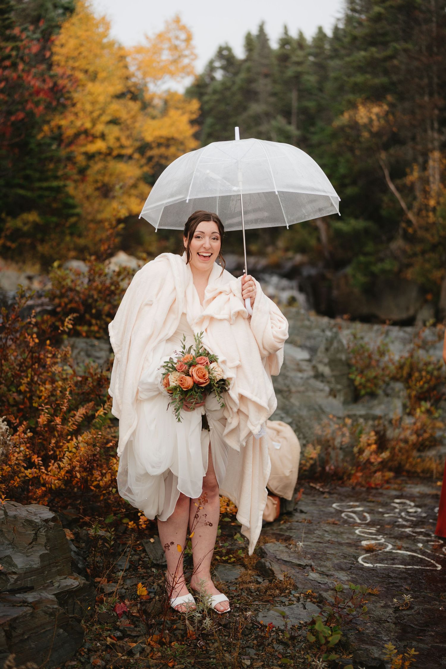 A bride in a white dress holds a clear umbrella while sitting on rocky terrain surrounded by autumn foliage.
