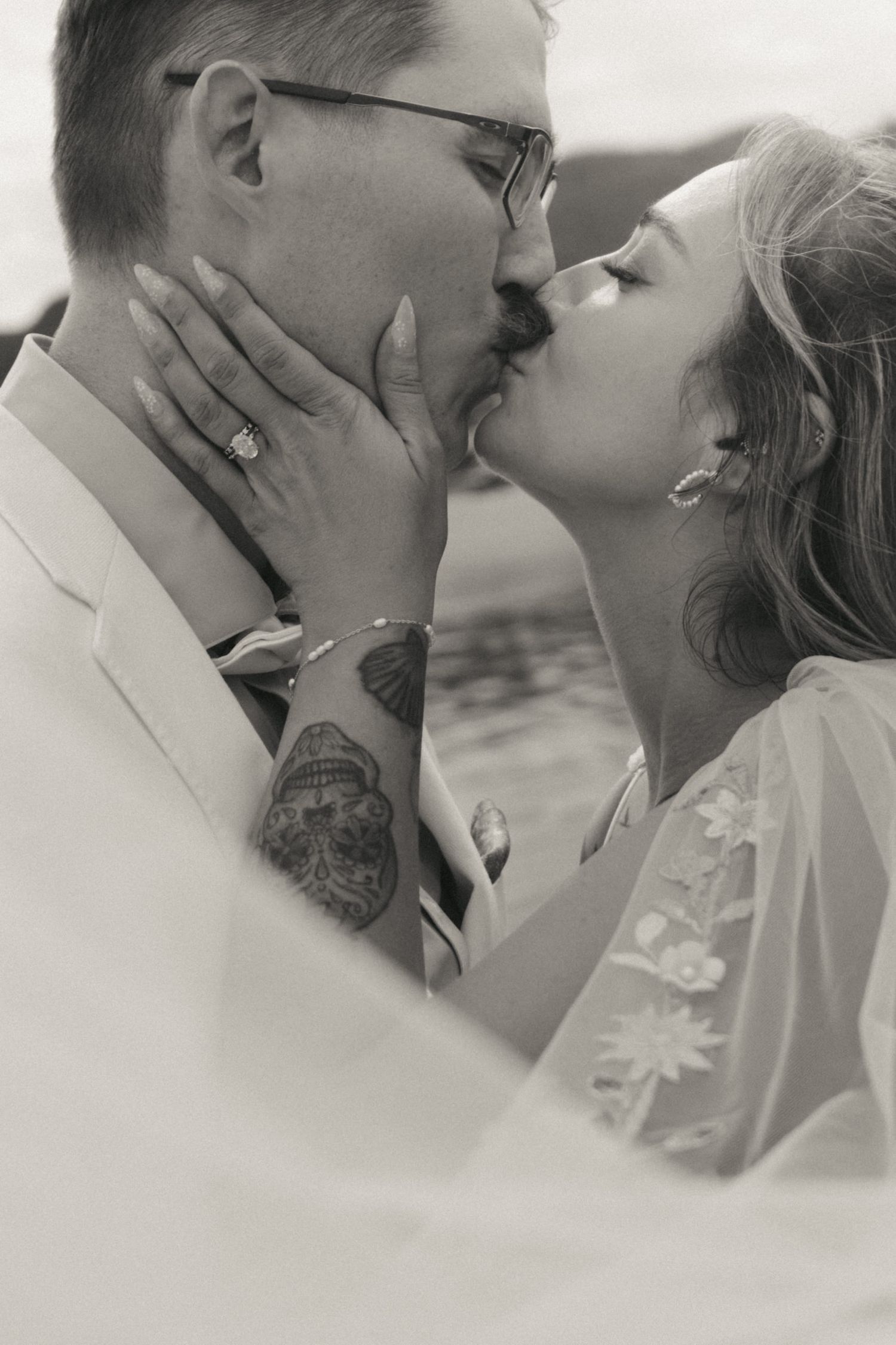 Black and white photo of a bride and groom sharing a romantic kiss, groom with tattoos and glasses wearing a white suit.