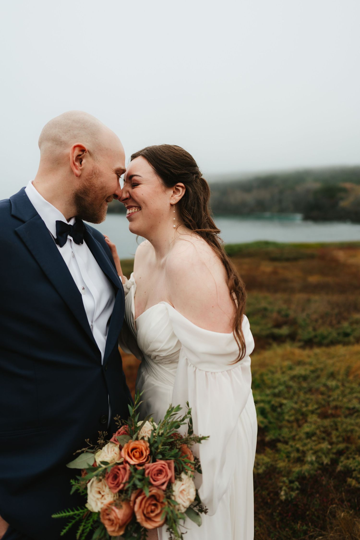 Couple sharing an intimate moment on a coastal cliffside with a peach and pink floral bouquet.