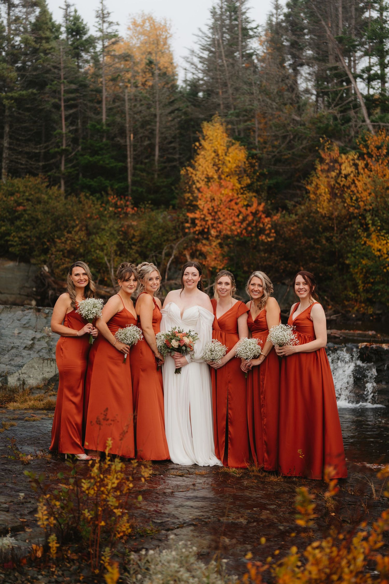 Wedding party in burgundy dresses stands by a waterfall surrounded by vibrant autumn foliage in the forest.
