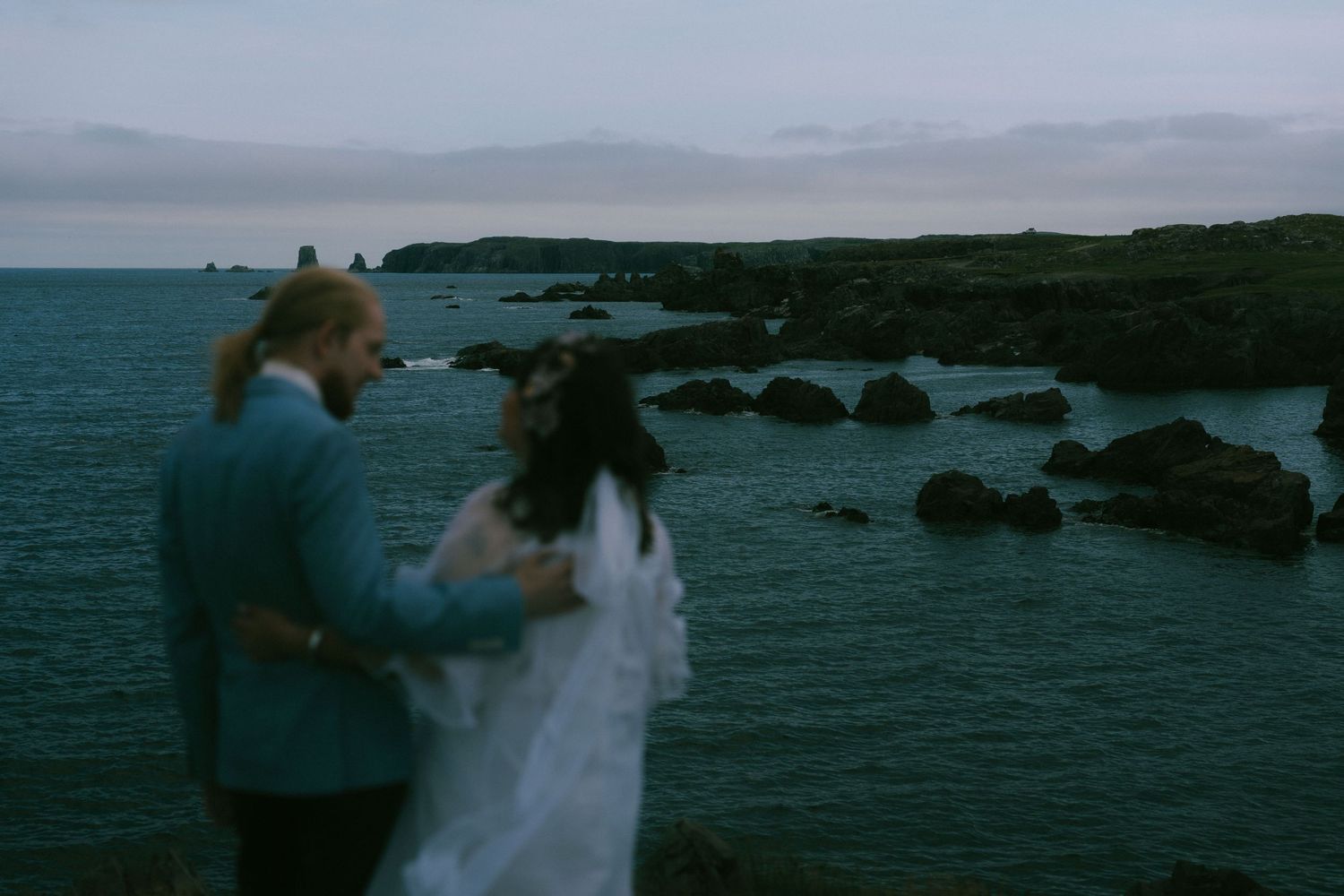 A couple walks along a scenic rocky coastline at sunset with ocean waves crashing against the shore.