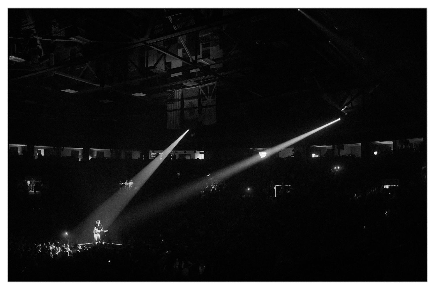 Dramatic spotlights illuminate a dark concert stage with silhouettes of performers and equipment visible in the shadows.