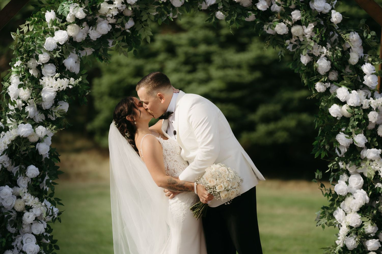 A romantic wedding couple shares a kiss under a beautiful floral arch in an outdoor garden setting.