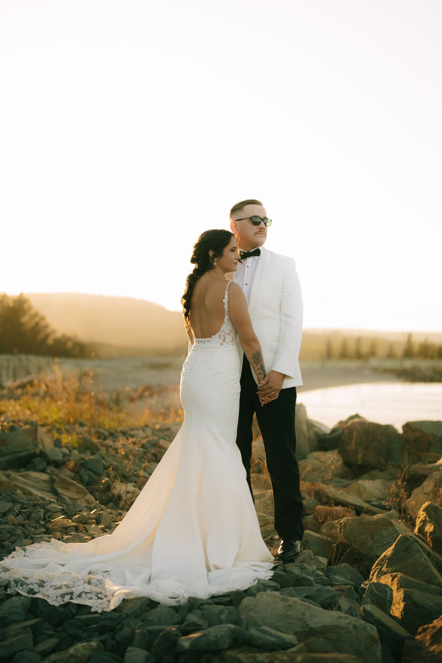 A couple in wedding attire poses together on rocky terrain during golden hour sunset by the water.