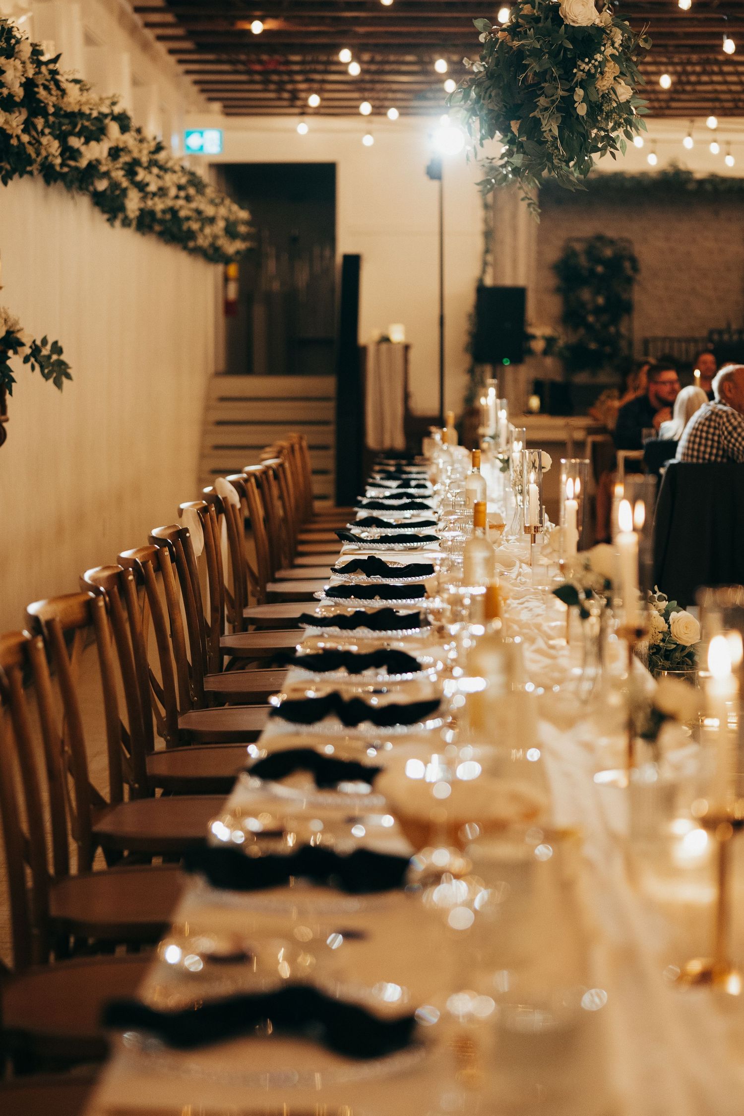 Long banquet table set for an elegant wedding reception with candlelight and greenery in an industrial-style venue.