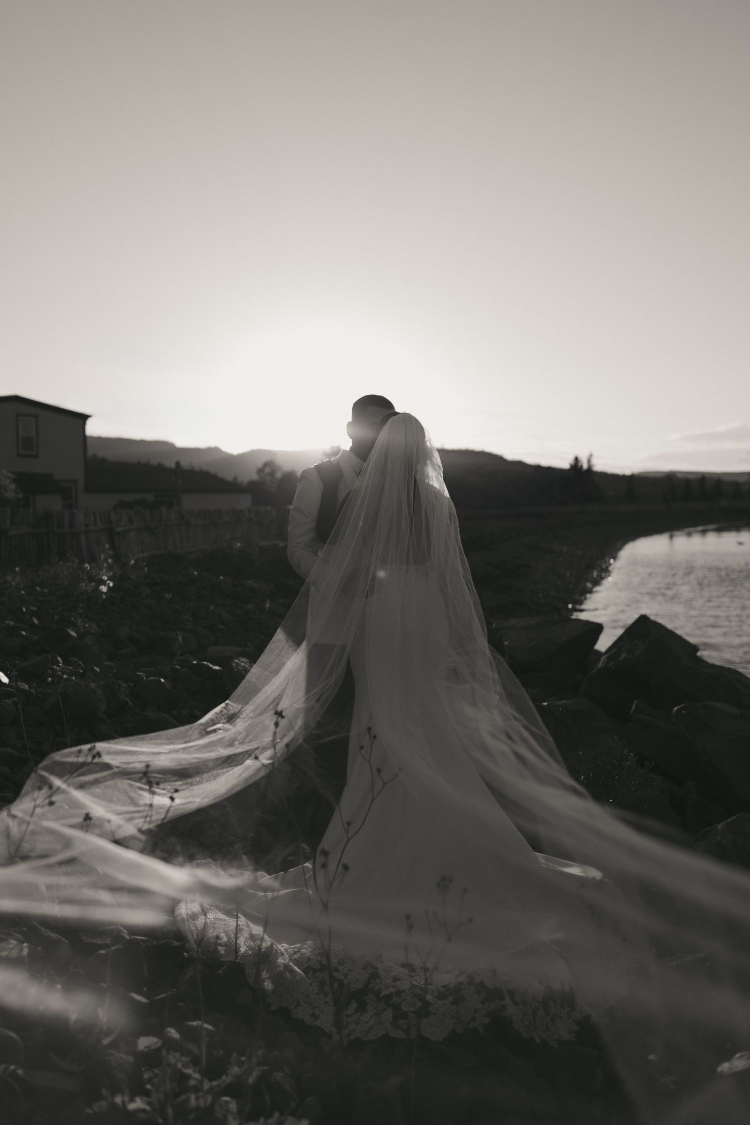 A bride in a flowing white veil stands silhouetted against a dramatic mountain landscape at sunset.