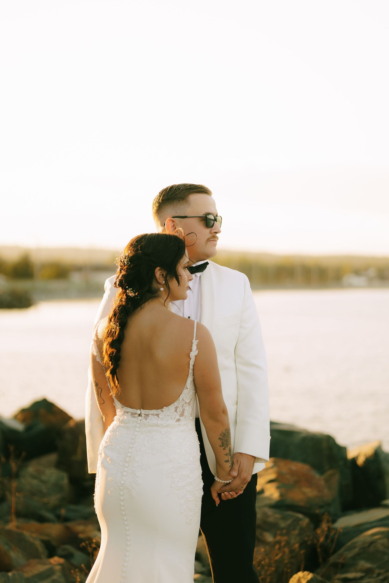 A couple in wedding attire holds hands at sunset on a rocky beach shoreline.