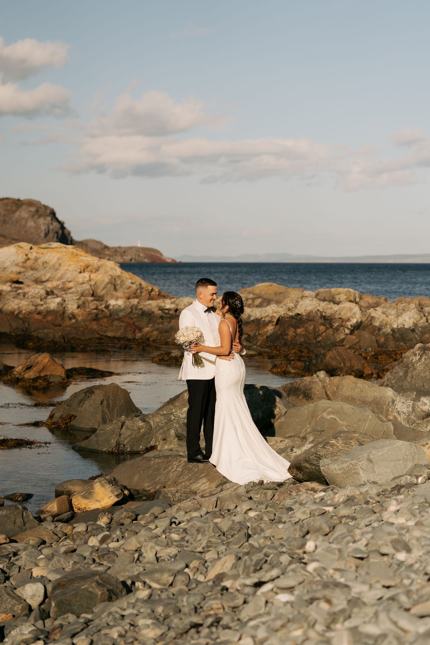 A romantic coastal photoshoot on rocky beach terrain with ocean and cloudy sky in the background during golden hour.