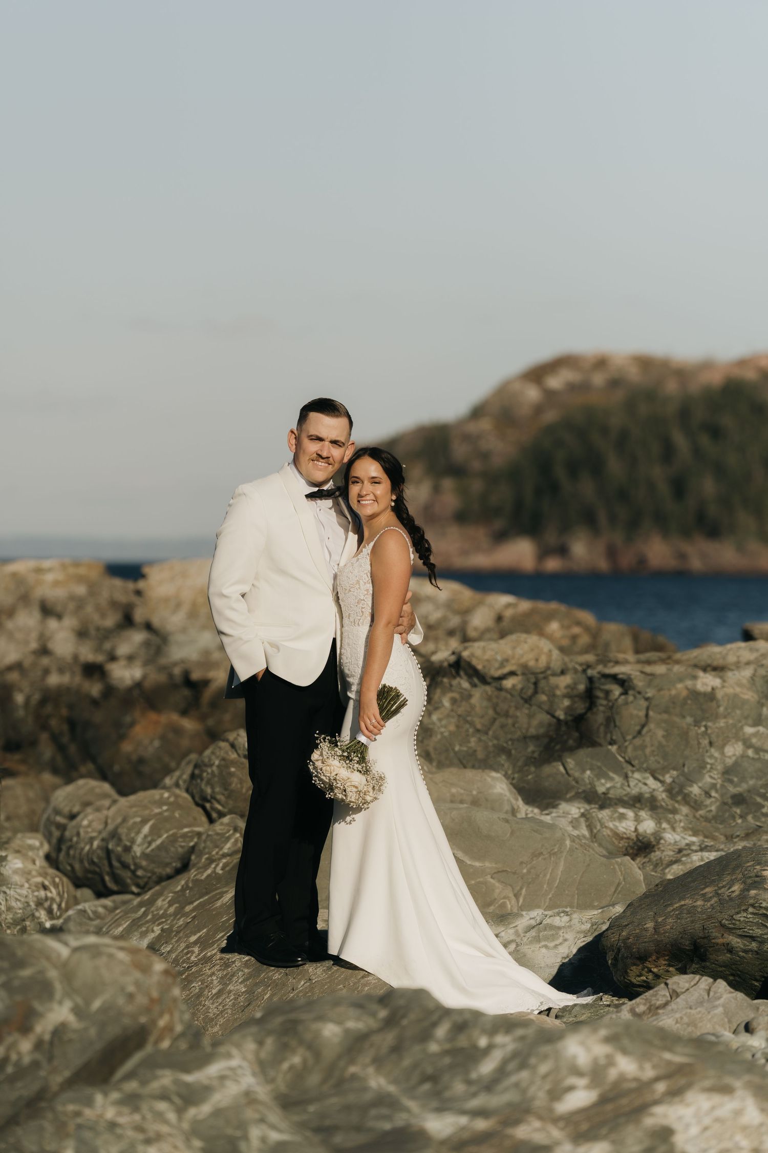 A couple in wedding attire embraces on rocky coastal cliffs with ocean and mountains in the background at sunset.