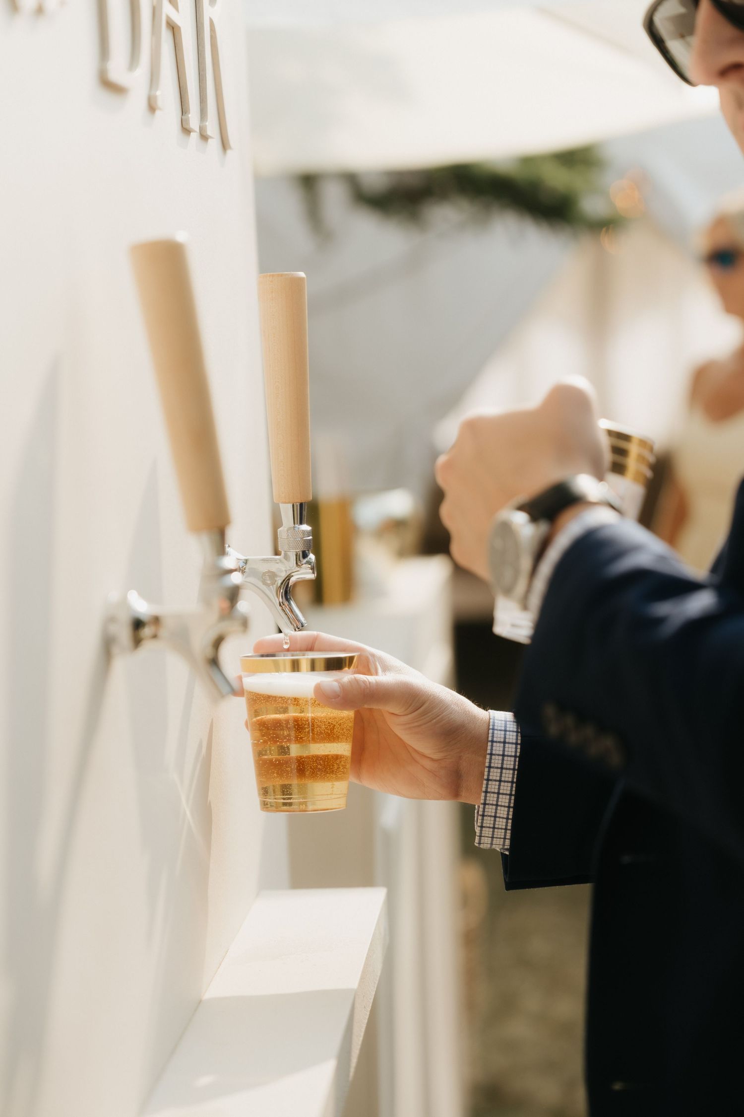 Hand holding a glass of beer through a white drink service window at an outdoor party.