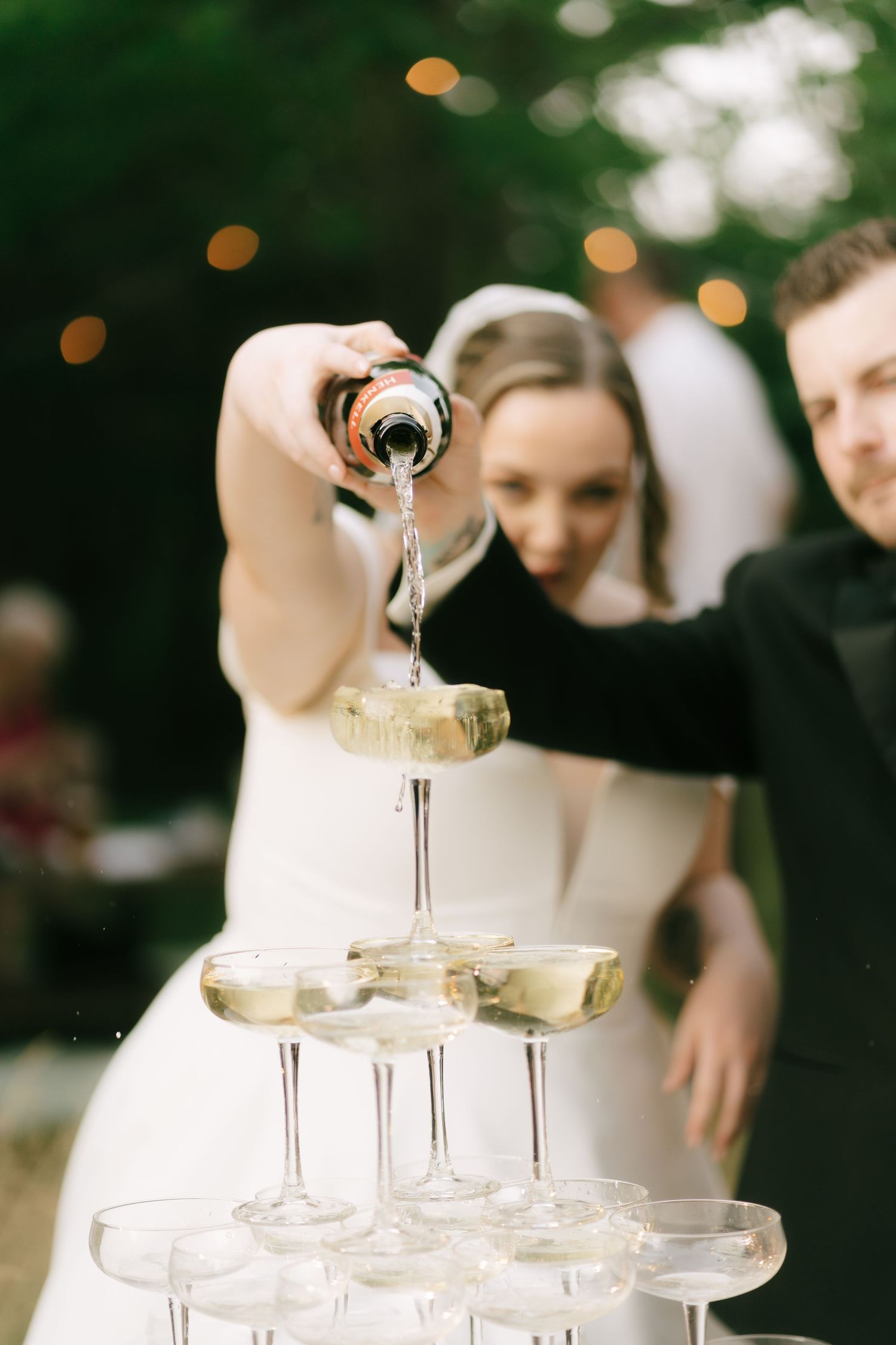 Champagne being poured into a tower of stacked coupe glasses at an outdoor celebration.