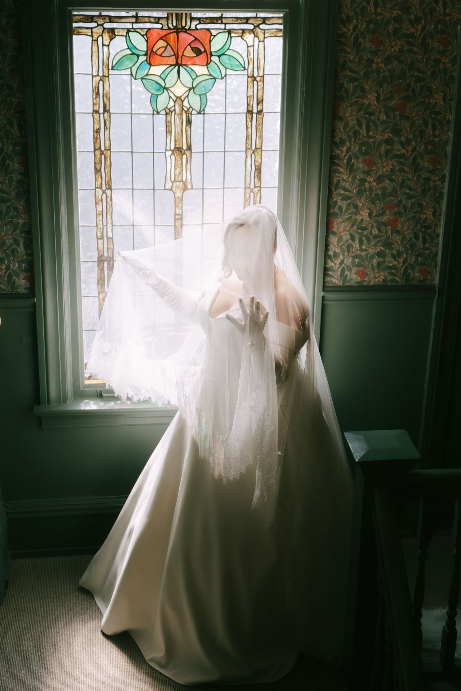 Wedding dress silhouetted against stained glass window with floral pattern and light streaming through.