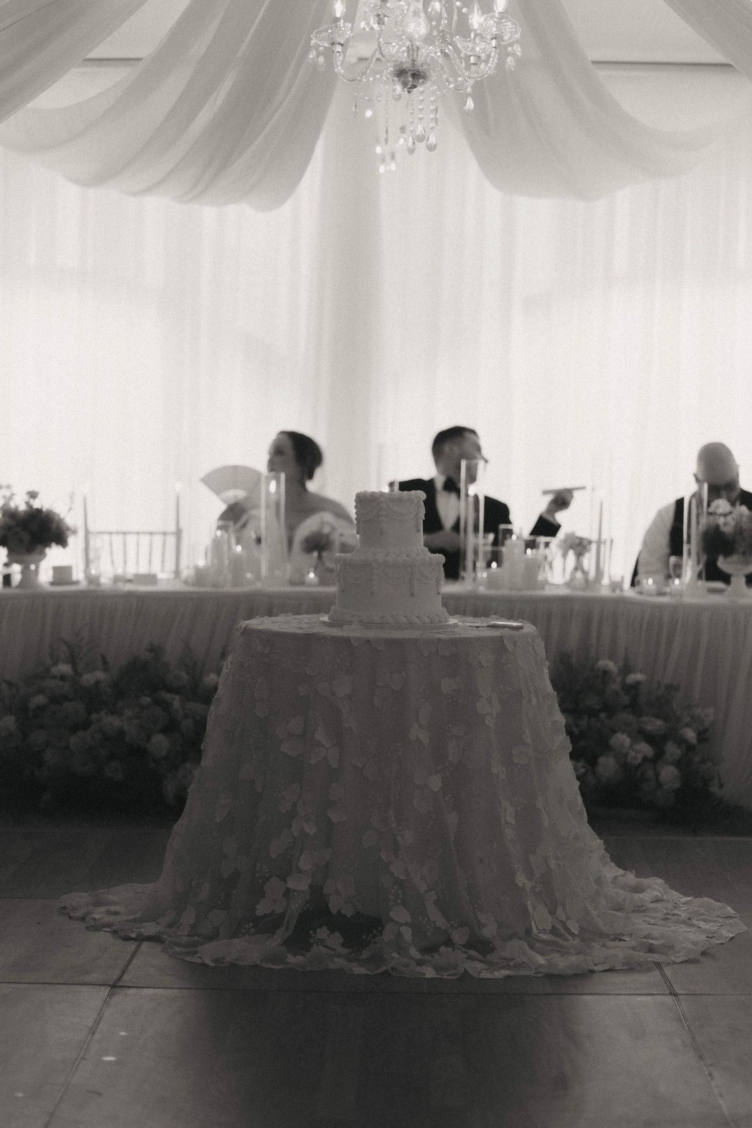 A black and white photo shows an elegant wedding reception setup with a chandelier and draped fabric ceiling.