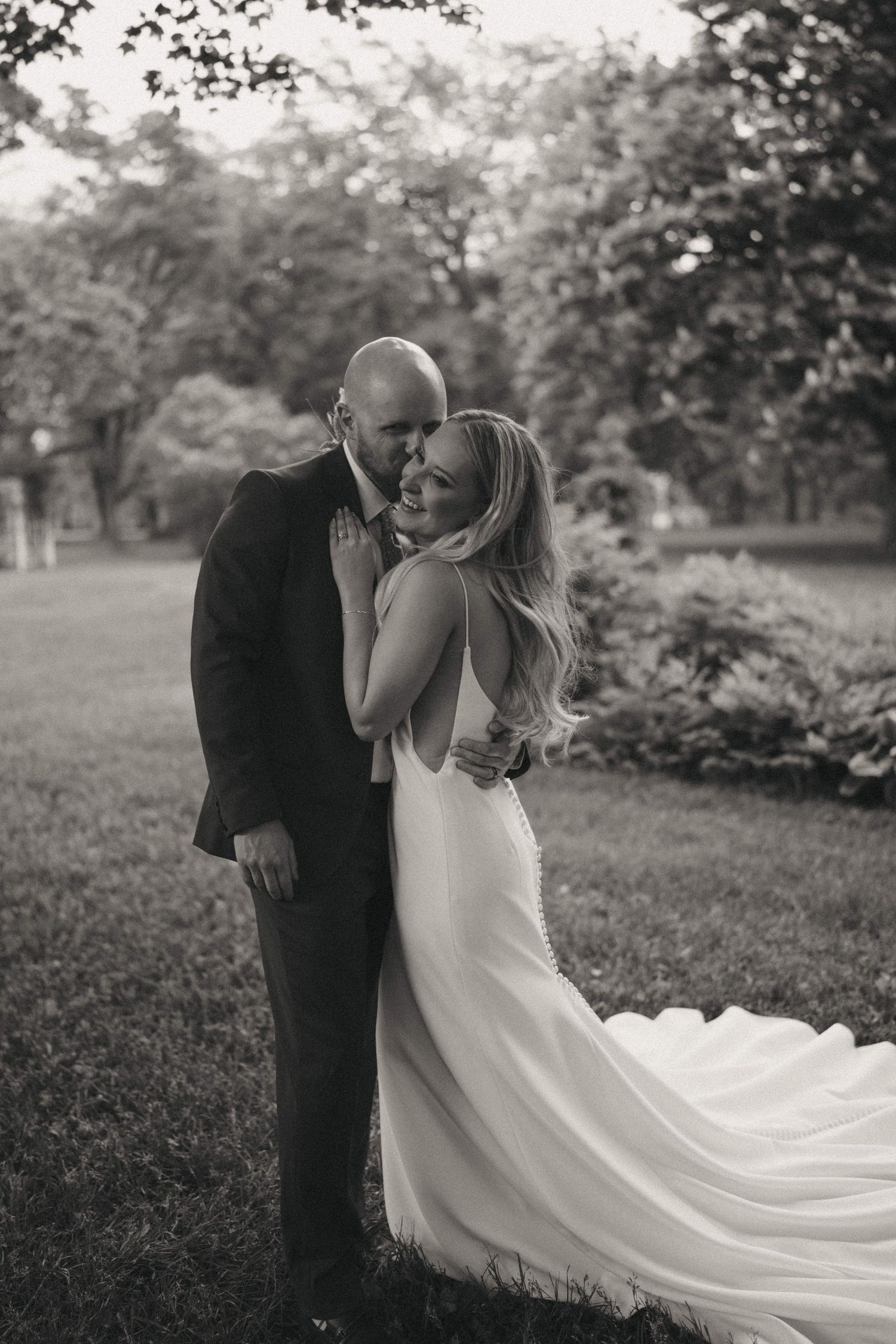 A flowing white wedding dress trails behind a couple embracing in a garden setting.