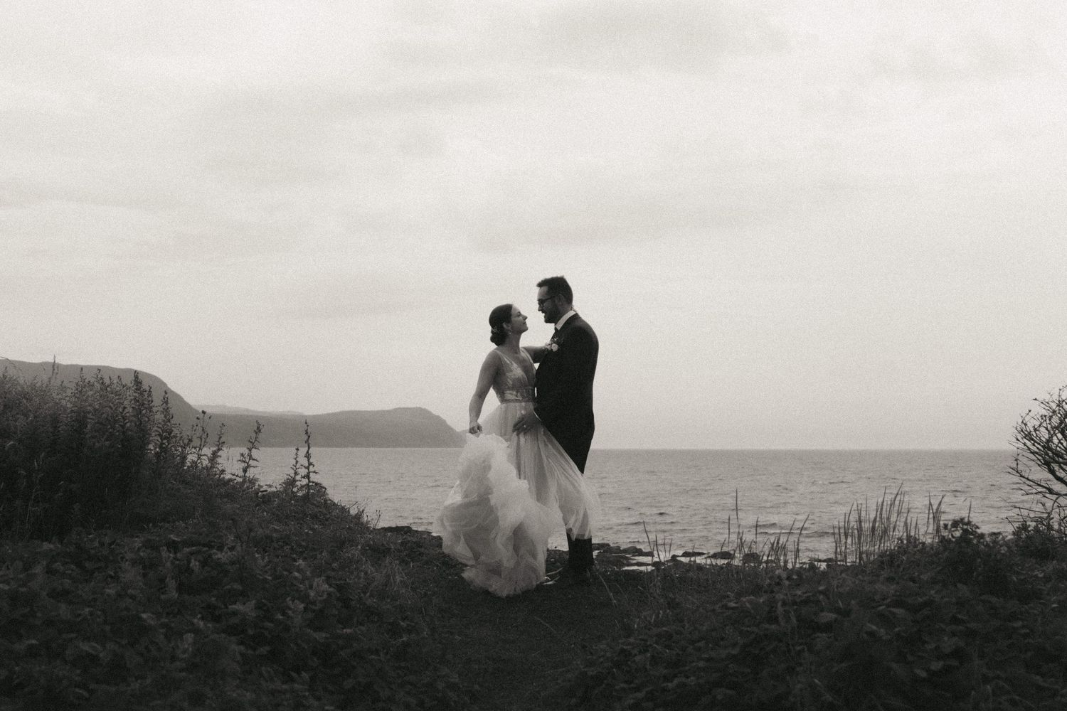 Silhouetted figures stand against a misty coastal landscape with rolling hills in the distance.