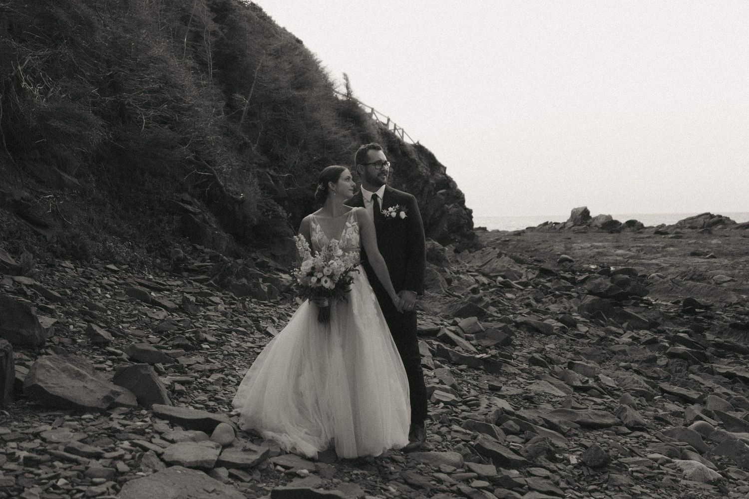 A couple in wedding attire walks along a dramatic rocky mountain path in black and white.