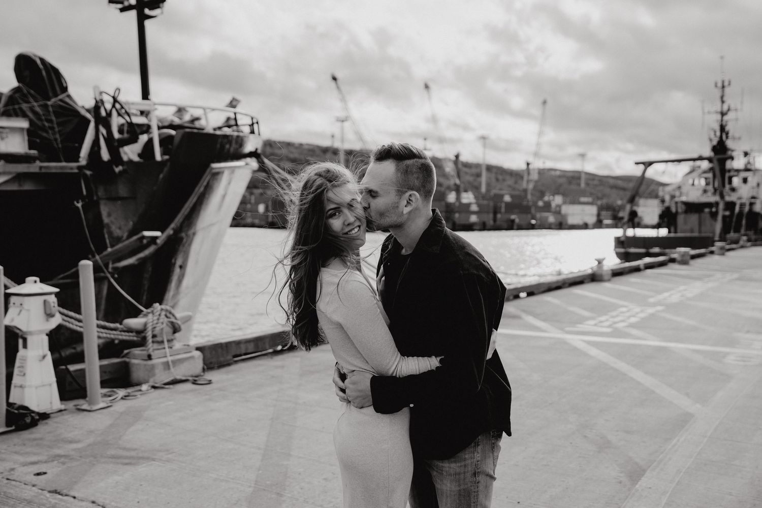 A tender moment between two people on an industrial dock with ships in the background.