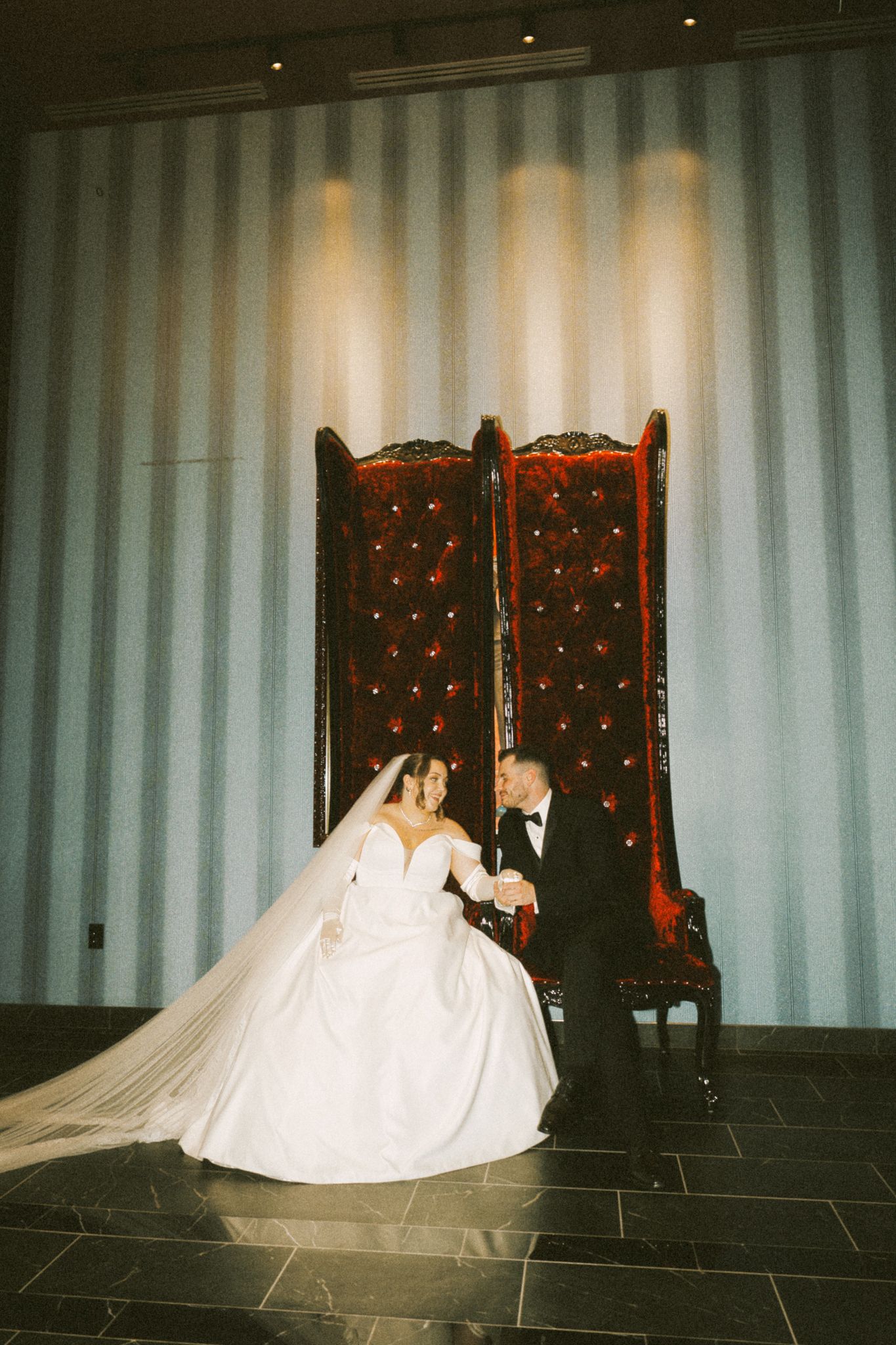 Wedding couple poses by ornate throne chairs against light backdrop.