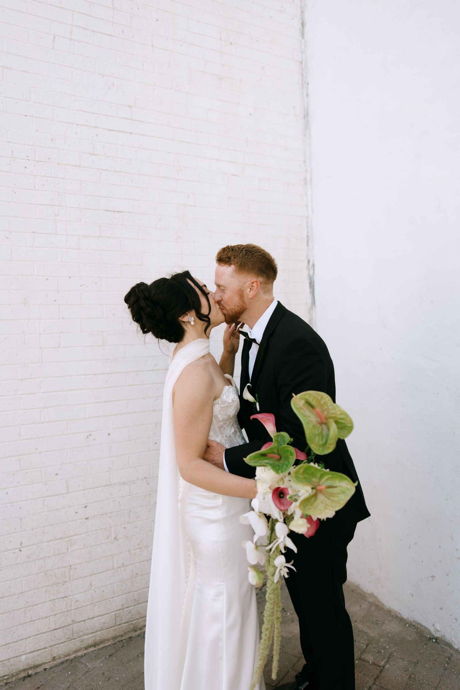 Bride and groom share a kiss against a white brick wall, bride holding a bouquet of tropical flowers.