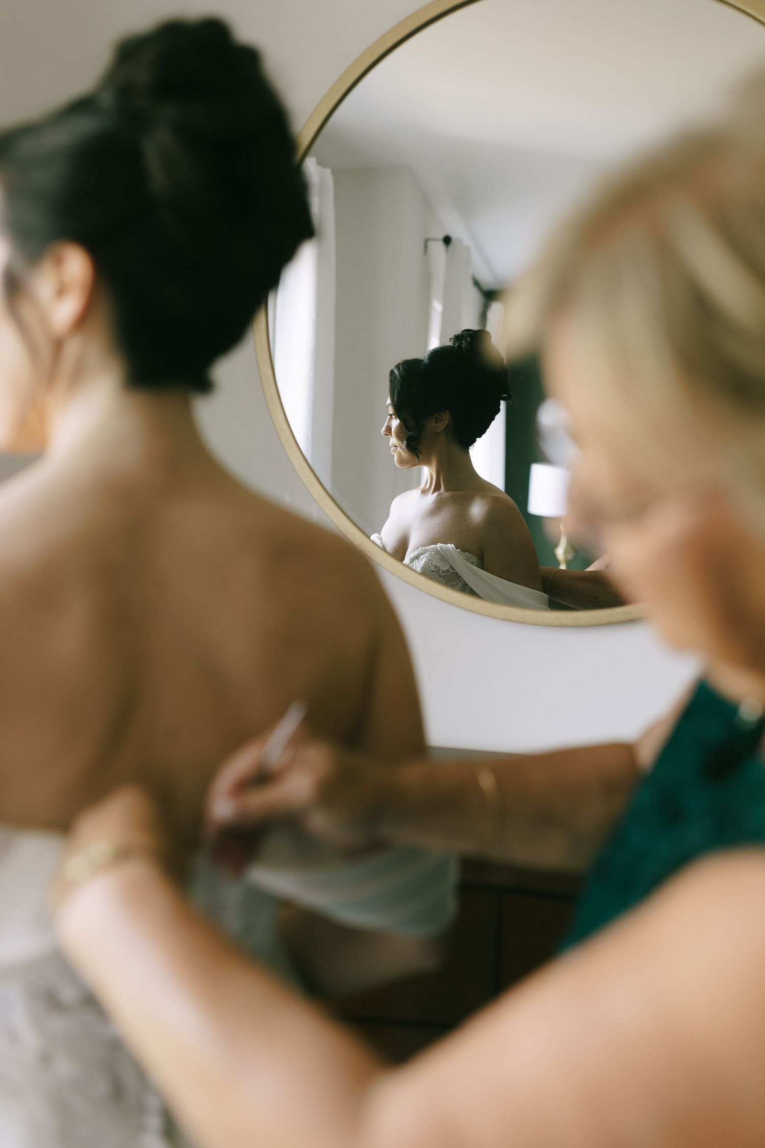 A bridesmaid helps a bride with her wedding dress while her reflection is visible in a round gold mirror.