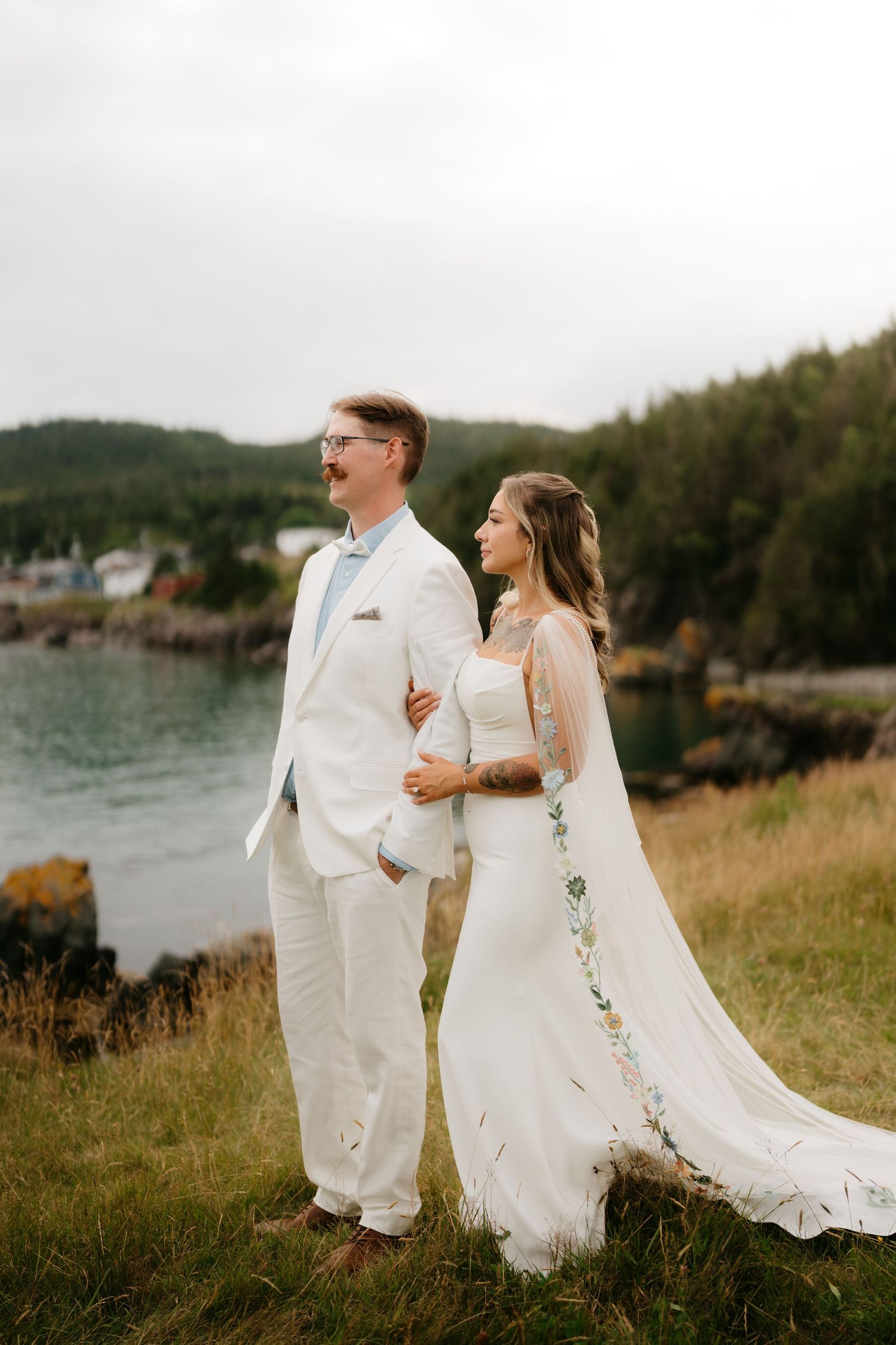 Bride and groom in white attire stand together on a grassy cliff overlooking a scenic coastal harbor.