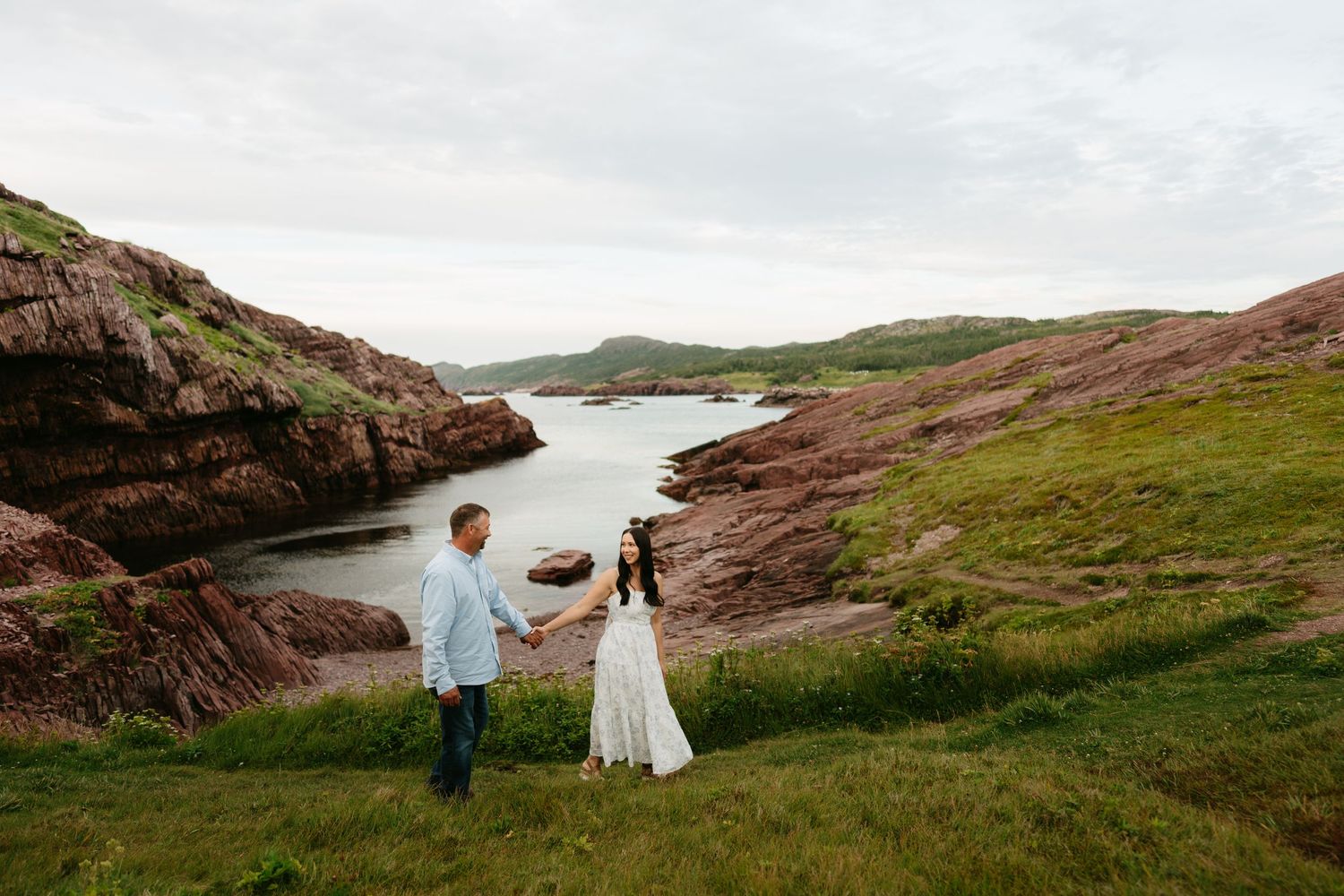 Couple holds hands on green cliffs above a rocky coastal inlet with red stone formations and calm water.