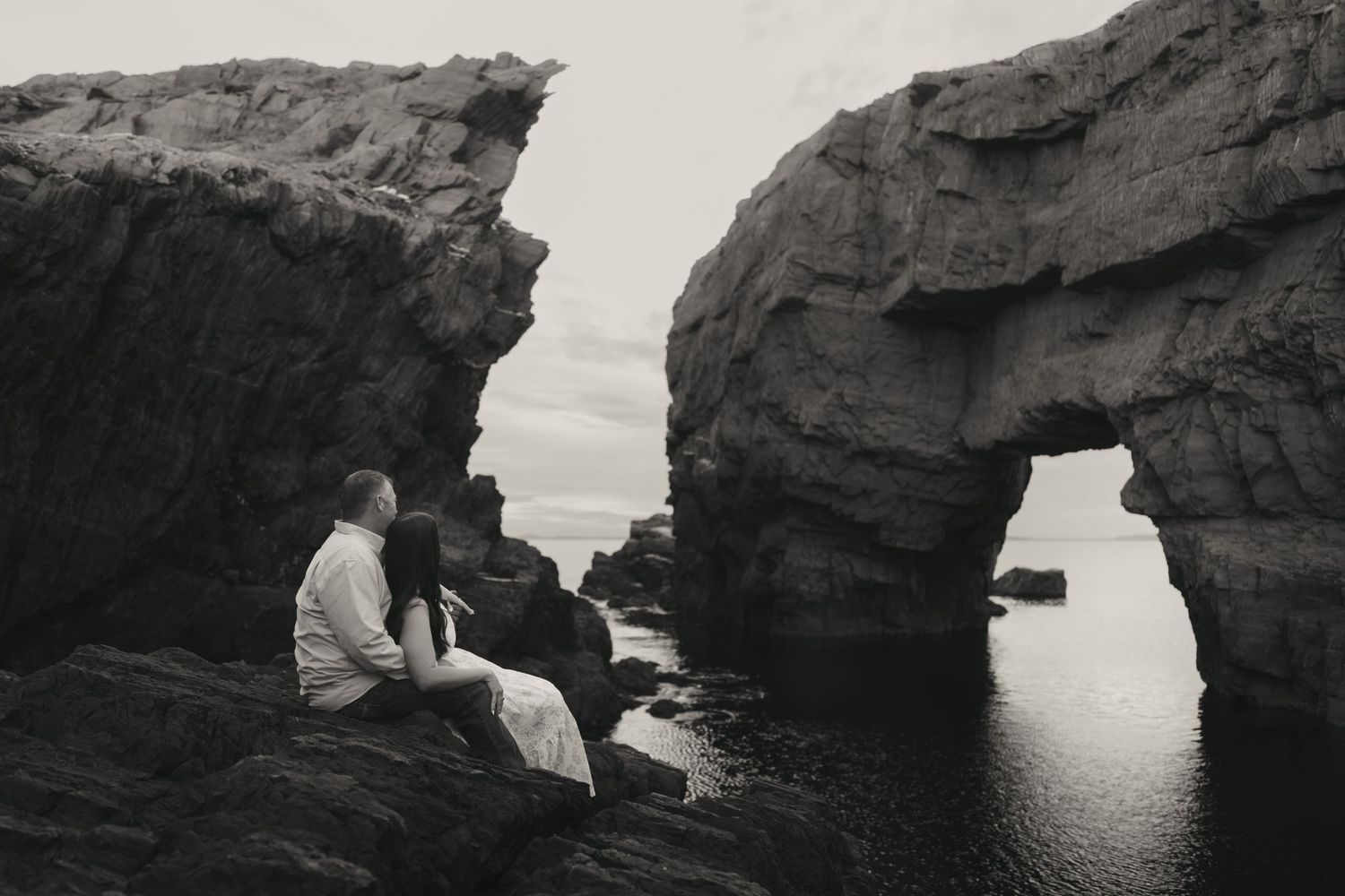 Couple sitting on rocky coastal cliffs beside a natural stone arch over calm ocean waters in black and white.